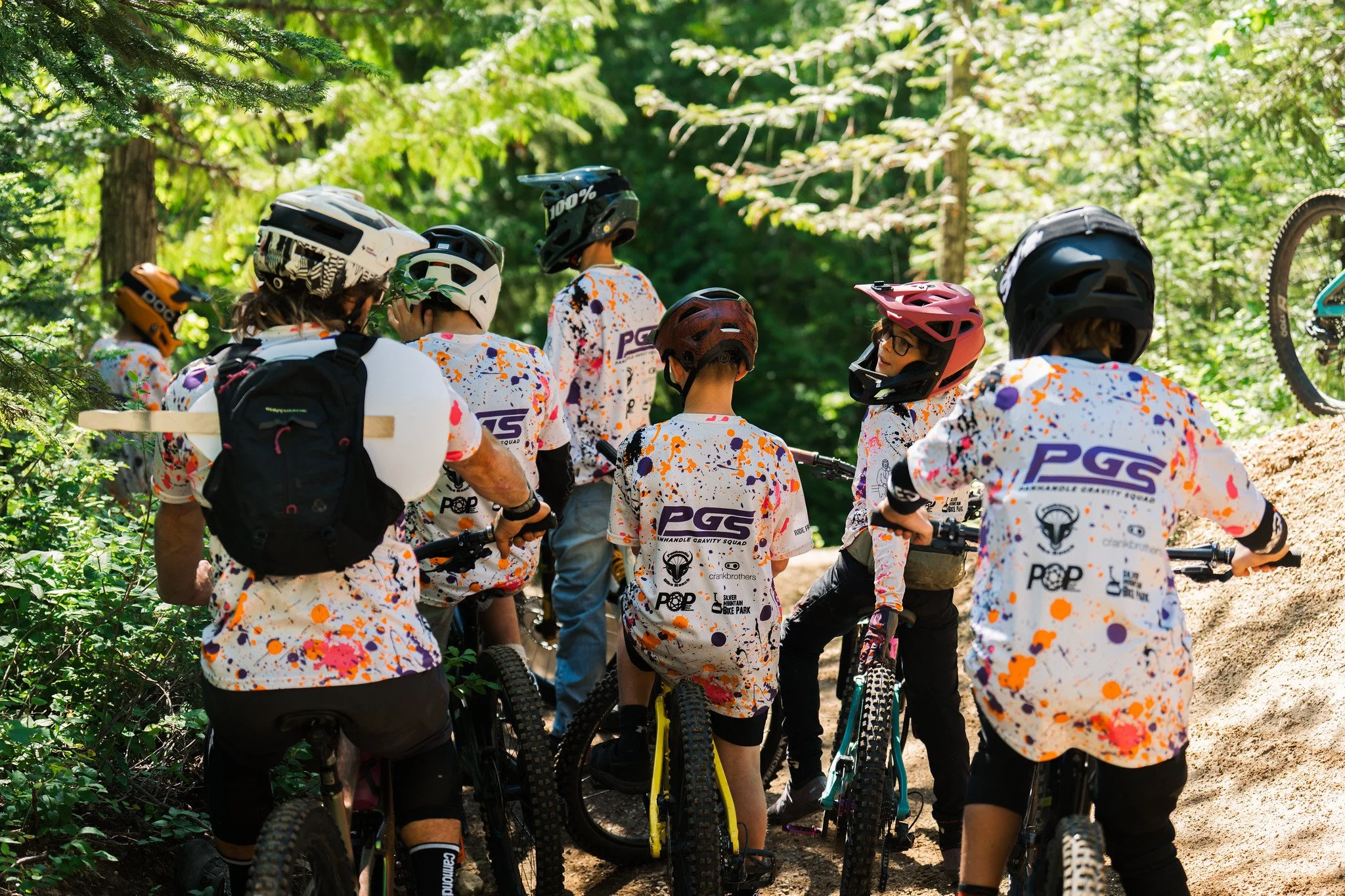 Group of mountain bikers wearing colorful jerseys and helmets, gathered with bikes on a forest trail.