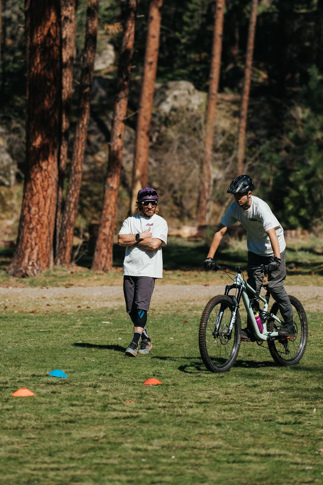 Two men outdoors on a grassy field, one is on a bicycle and the other is standing with arms crossed, near small blue and orange cones.