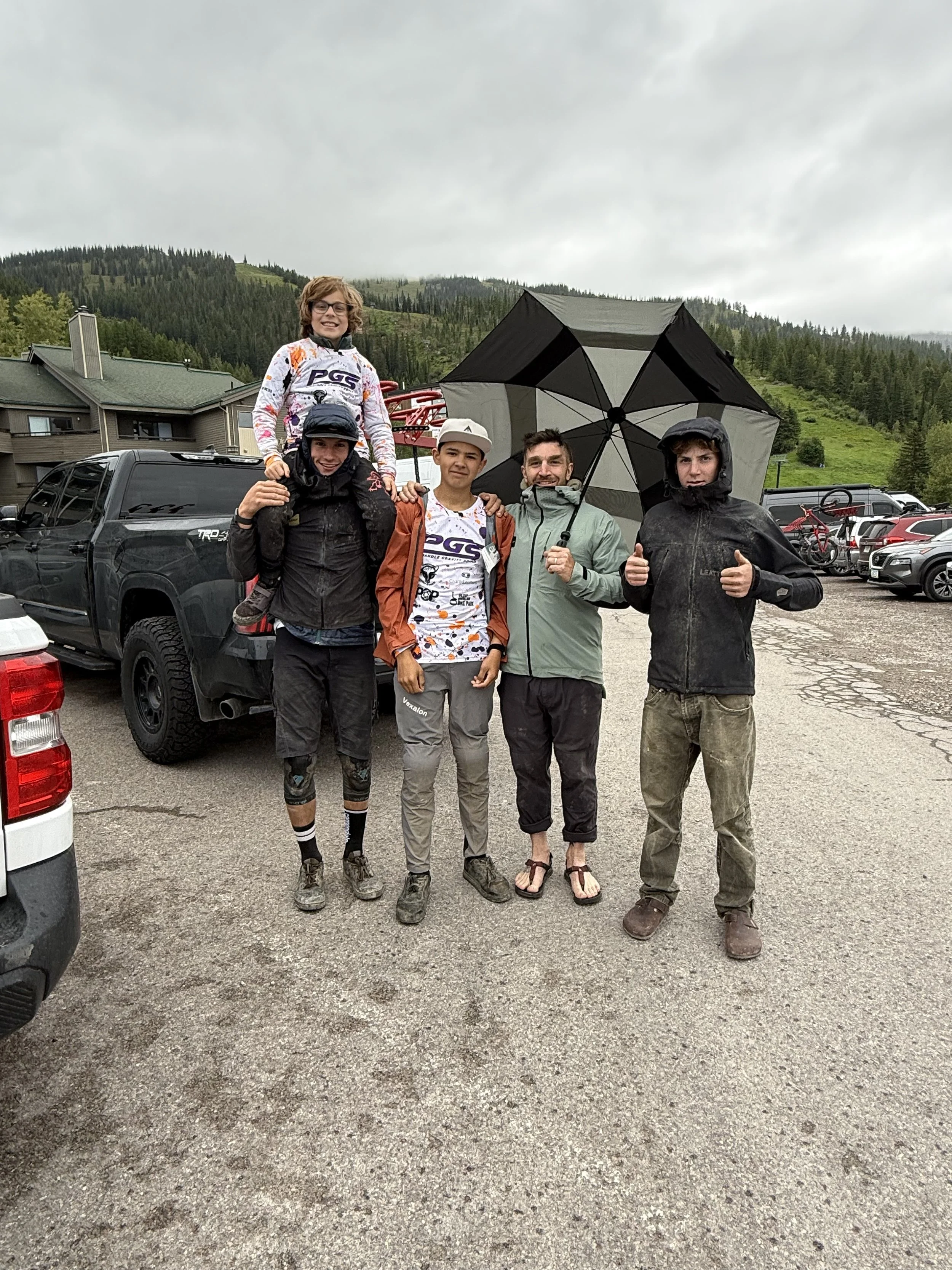A group of five people outdoors in a parking lot with a mountain and forest background, holding an umbrella, smiling and wearing casual and mountain biking clothing.