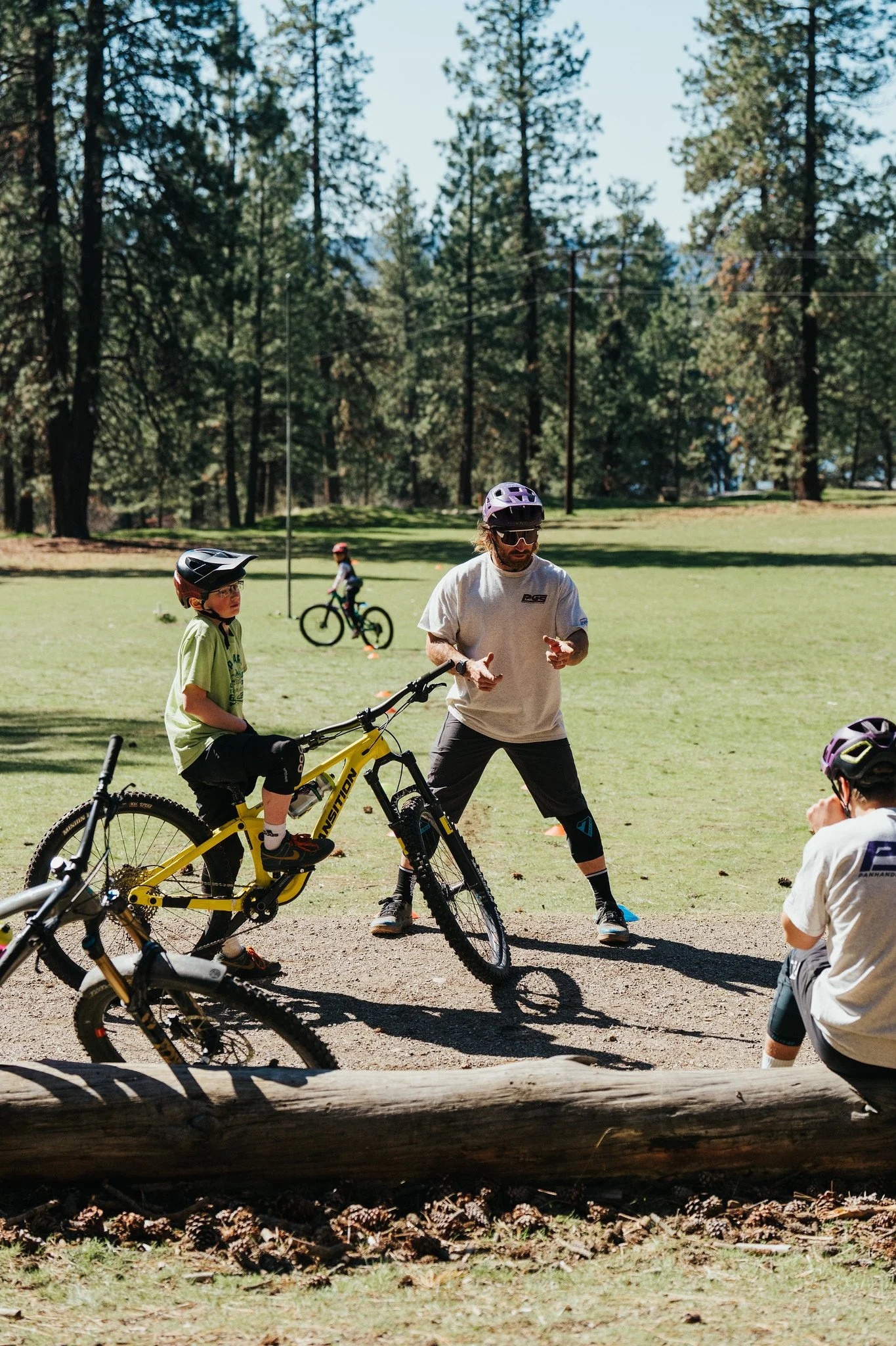 Children and an adult in a park, some children on bikes and an adult standing near a yellow mountain bike, all wearing helmets, with trees in the background.