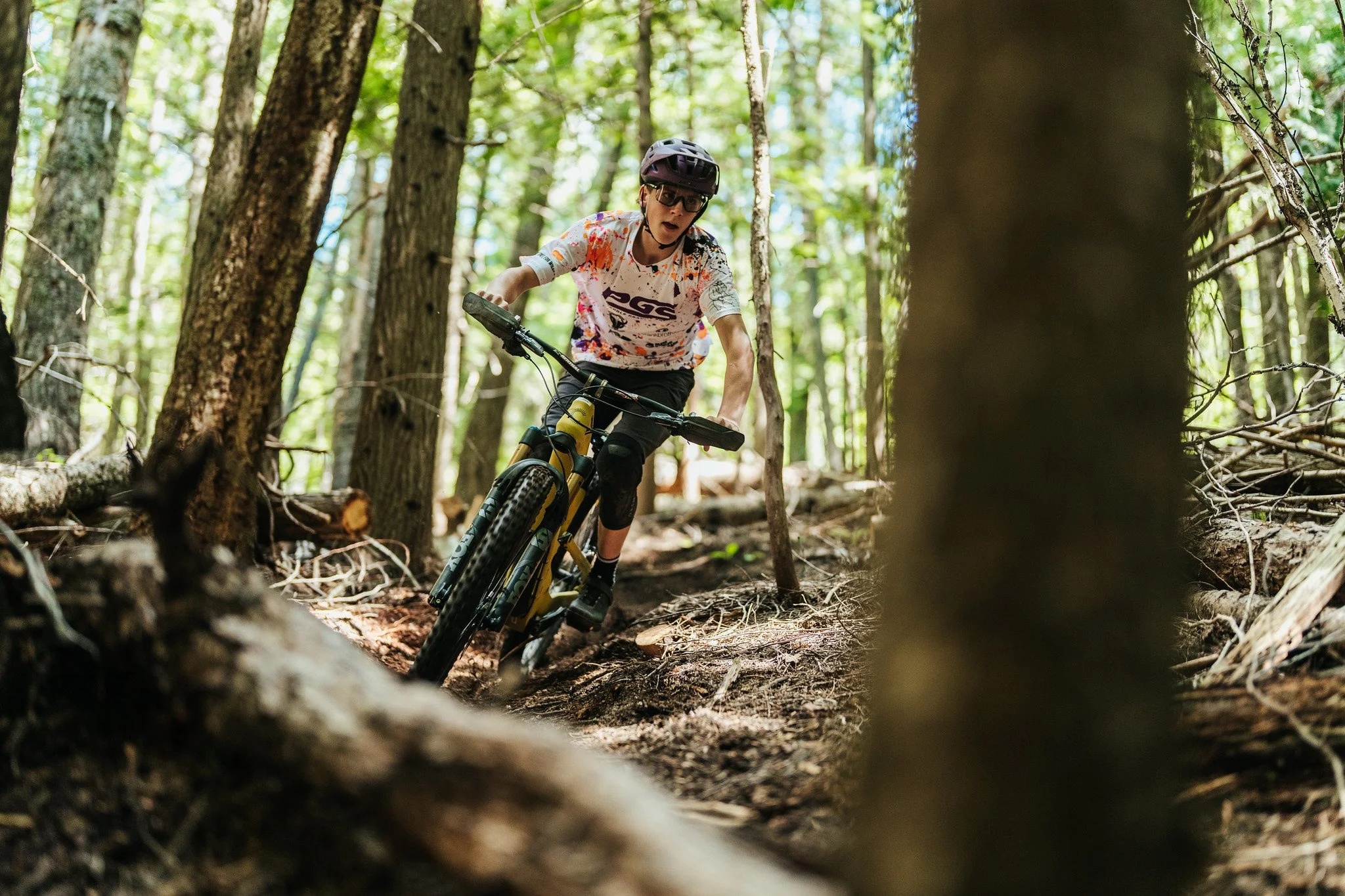 Person mountain biking through a dense forest, wearing a helmet and glasses, and a white and splattered shirt.