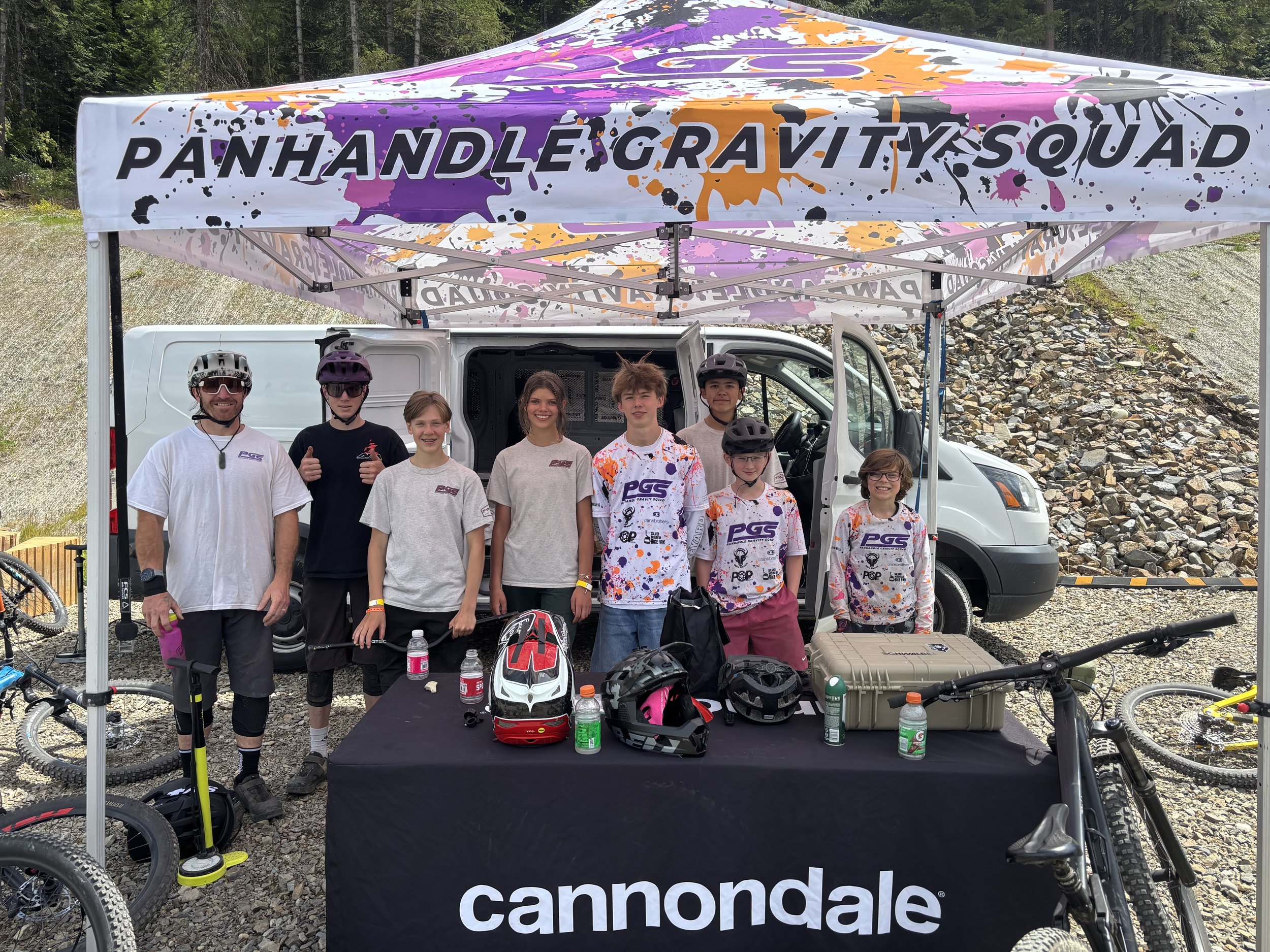 A group of seven people, including children and adults, standing under a colorful canopy tent with the words 'P&H Gravity Squad' on it. They are dressed in cycling gear, some wearing helmets, and there are bikes and cycling equipment on a table in fr