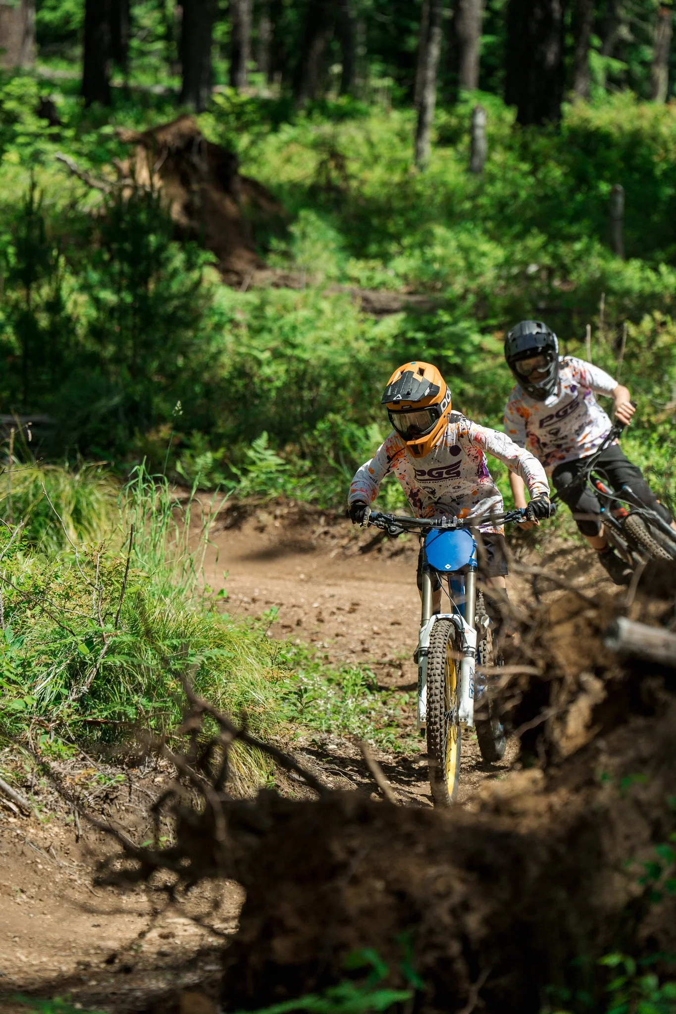 Two mountain bikers riding on a dirt trail through a dense green forest, with the riders wearing helmets and protective gear.