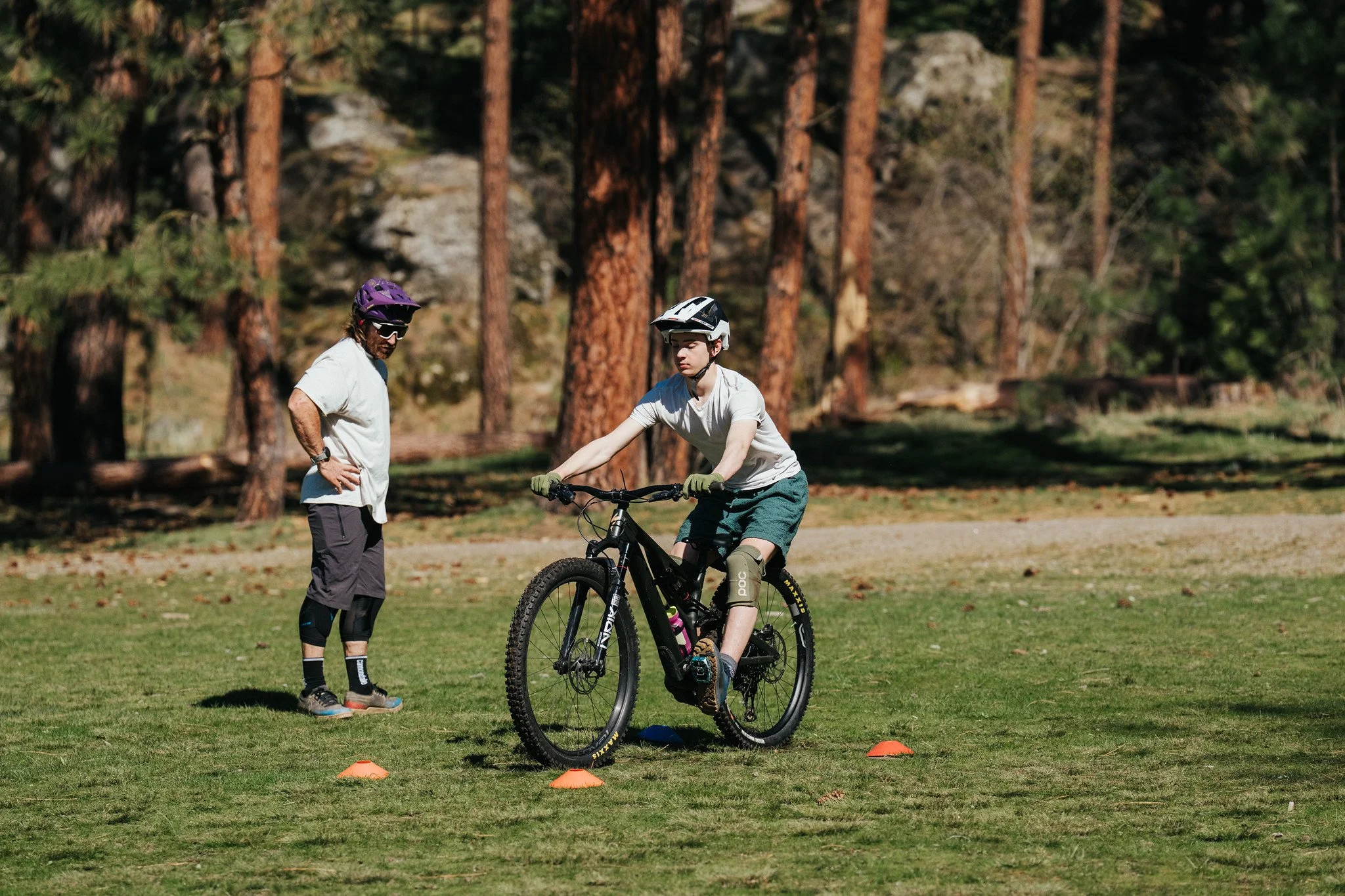 A boy wearing a helmet and gloves is learning to ride a mountain bike with an instructor or parent standing nearby in a park with tall trees.