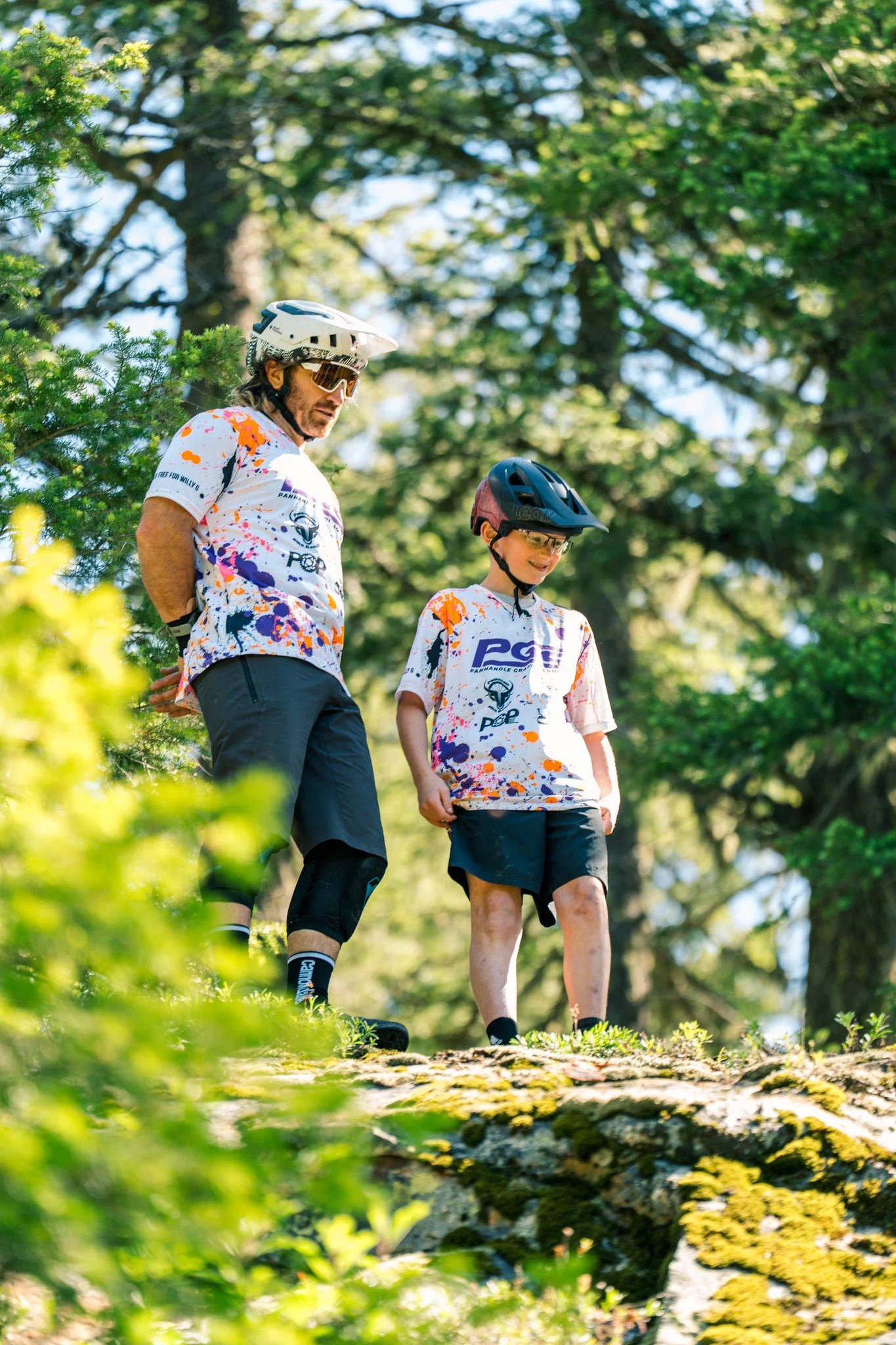 A man and a boy, both wearing sports jerseys, helmets, and safety gear, are walking outdoors in a lush, green forested area.