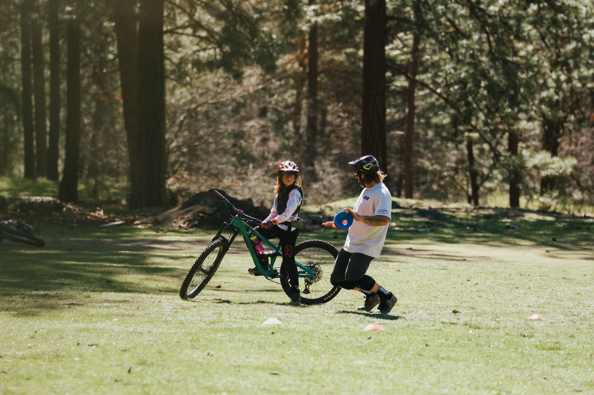 A young girl on a mountain bike wearing a helmet, being helped by a man in a park with tall trees, during daytime.