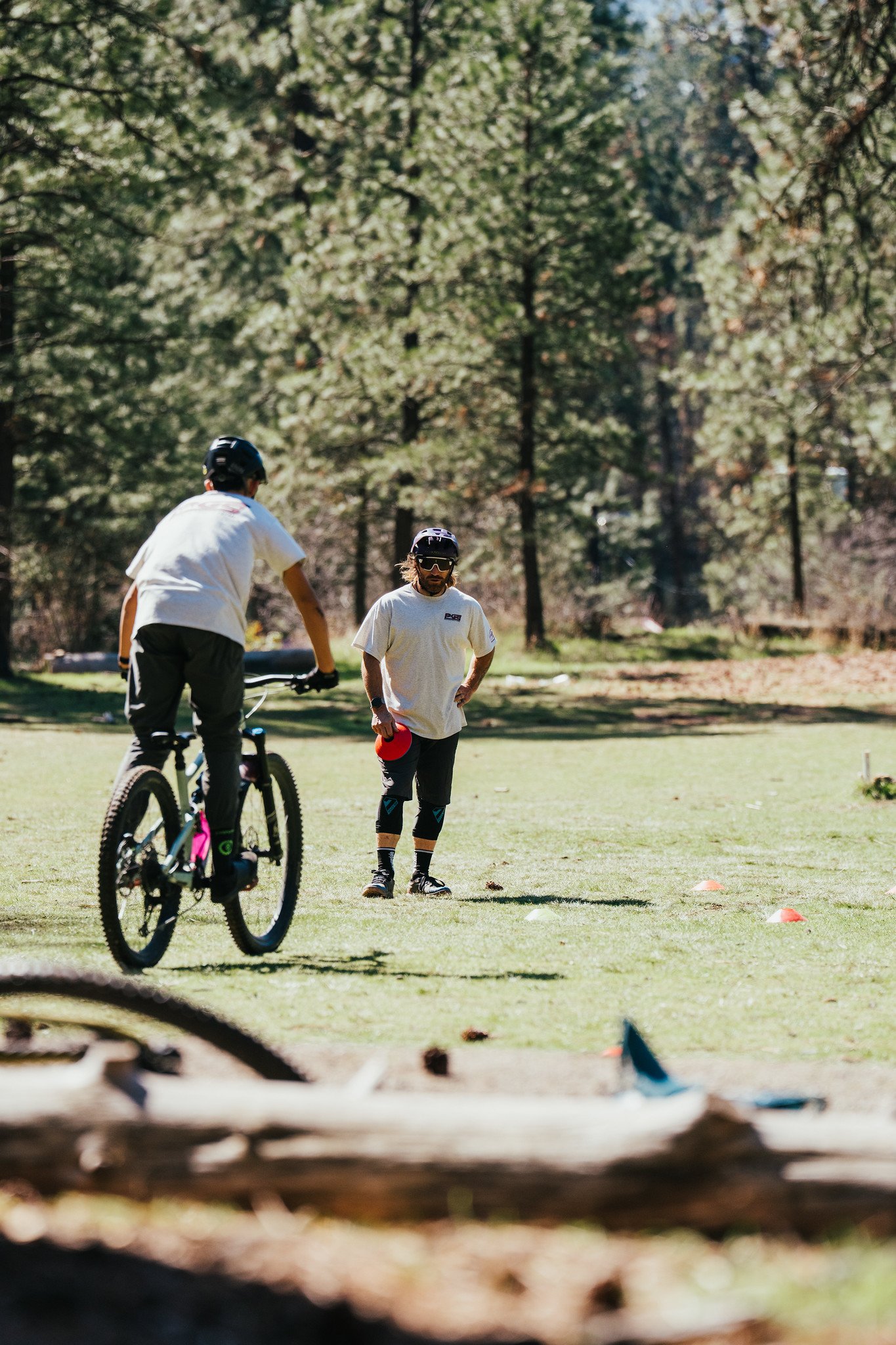 Two young men in helmets and casual clothing participating in an outdoor activity, with one riding a bicycle and the other standing, surrounded by trees in a park or wooded area.