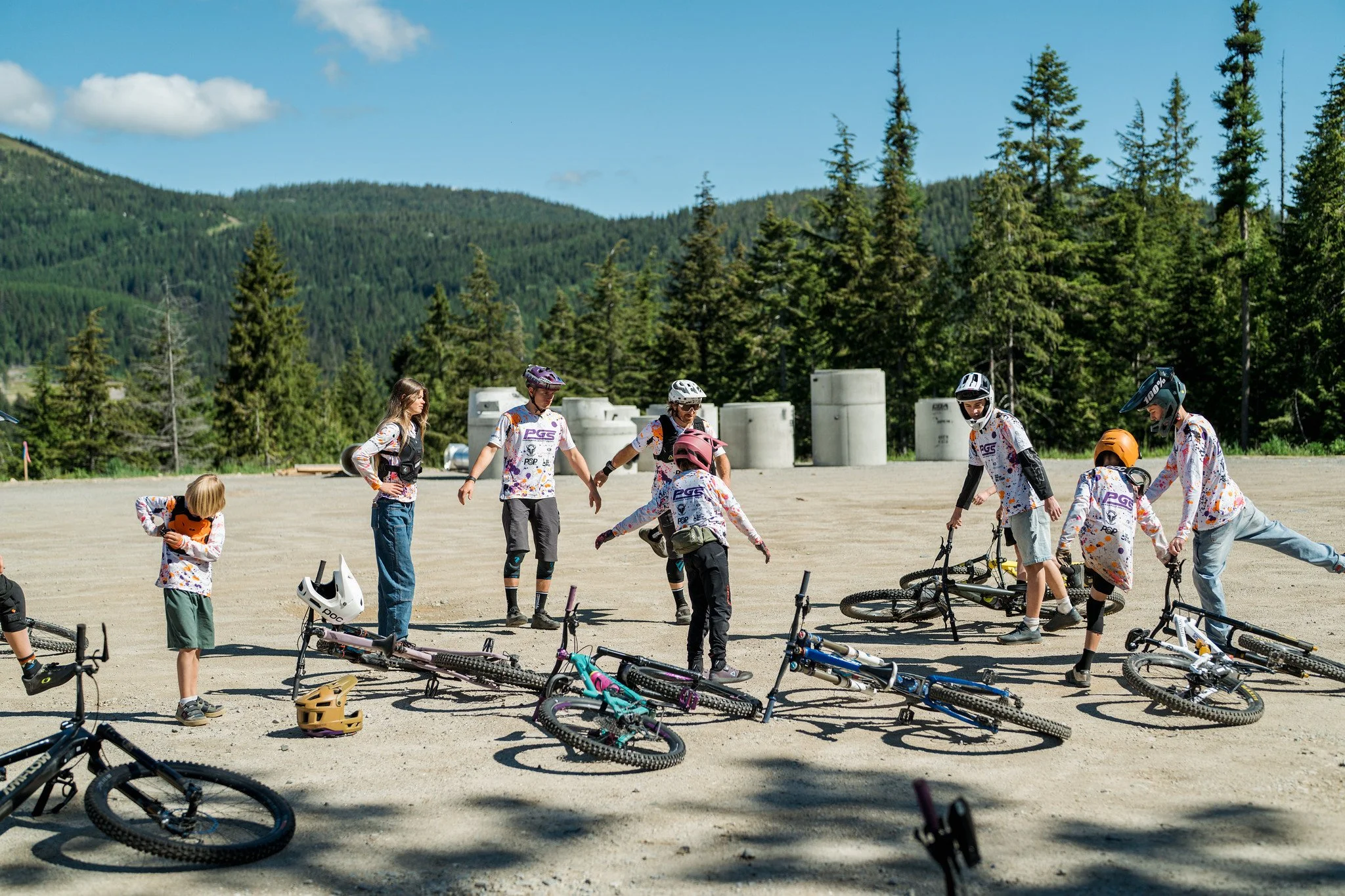 Group of children and teenagers in mountain biking gear, some with helmets, standing and sitting on the ground with their bikes, in a forested outdoor area under a blue sky.