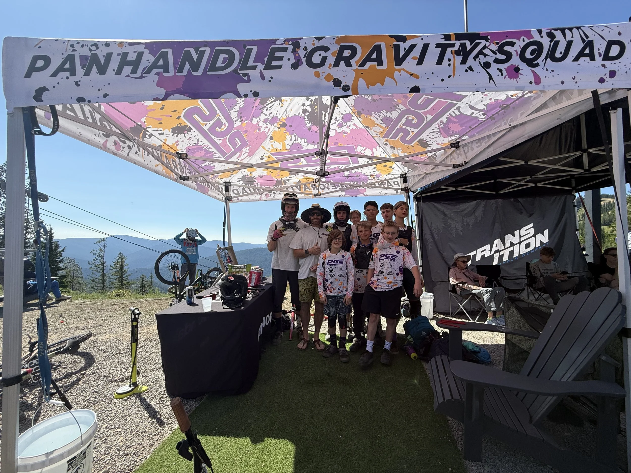 A group of young mountain bikers and their coaches are gathered under a colorful canopy at a mountain biking event, with bikes and event gear visible around them.
