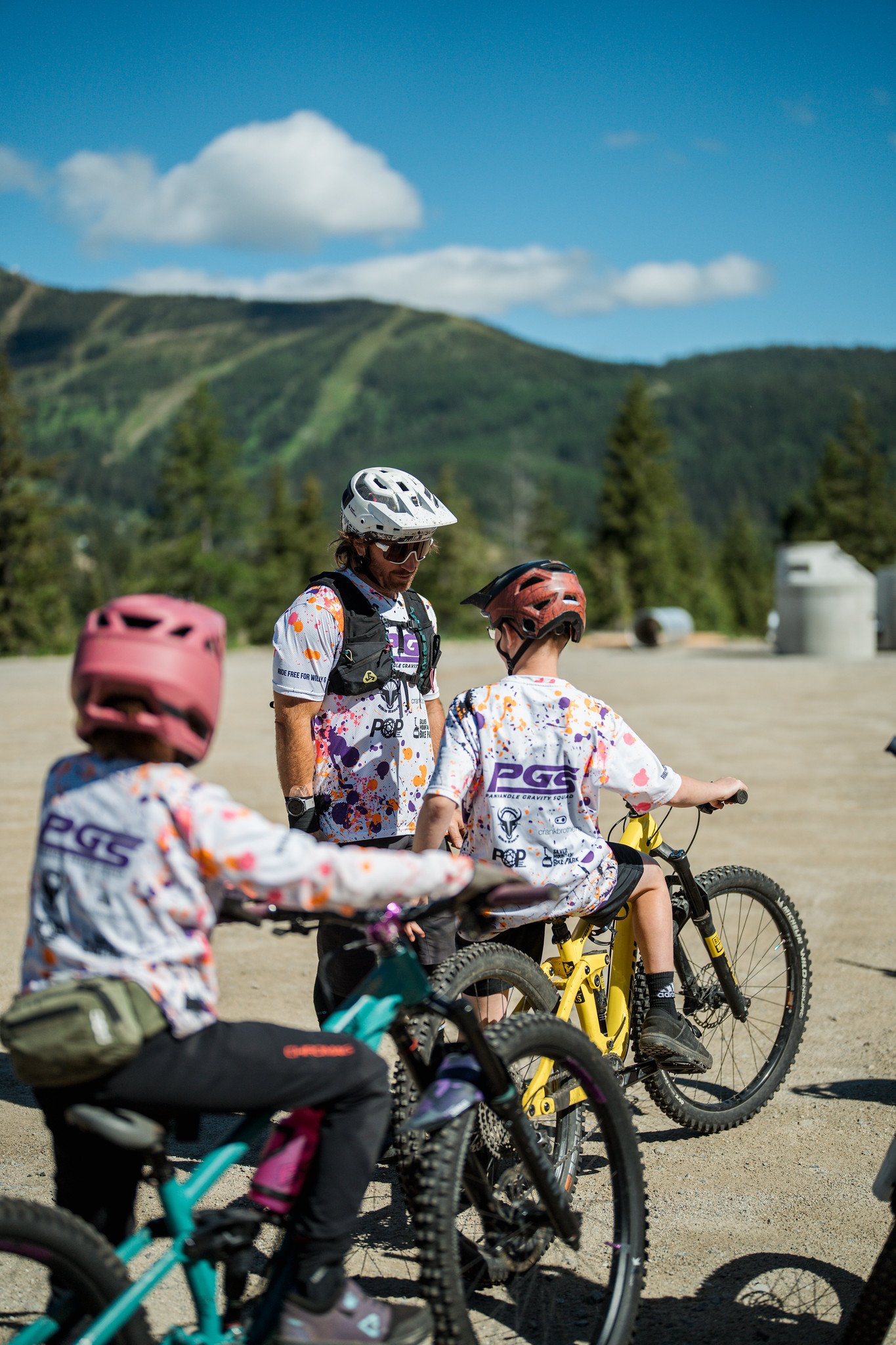 A group of young children wearing helmets and colorful shirts riding bikes outdoors, with a mountain range and trees in the background on a sunny day.