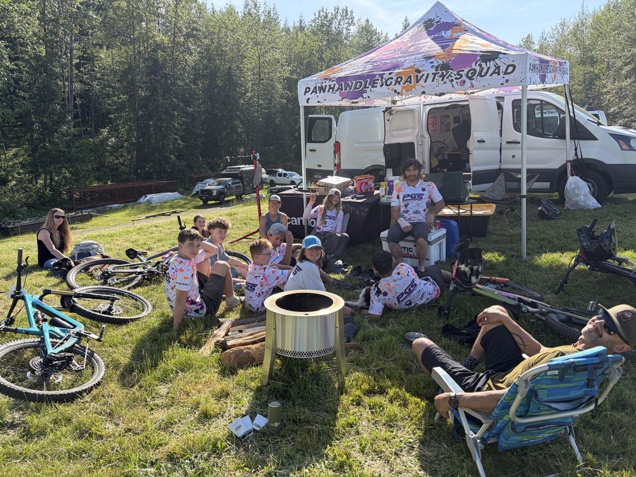 Group of people, including children and adults, sitting and relaxing outdoors on grass near a white van and a canopy tent that reads 'Panhandle Gravity Squad'. Bicycles are on the ground nearby, and the setting appears to be a sunny park or outdoor e