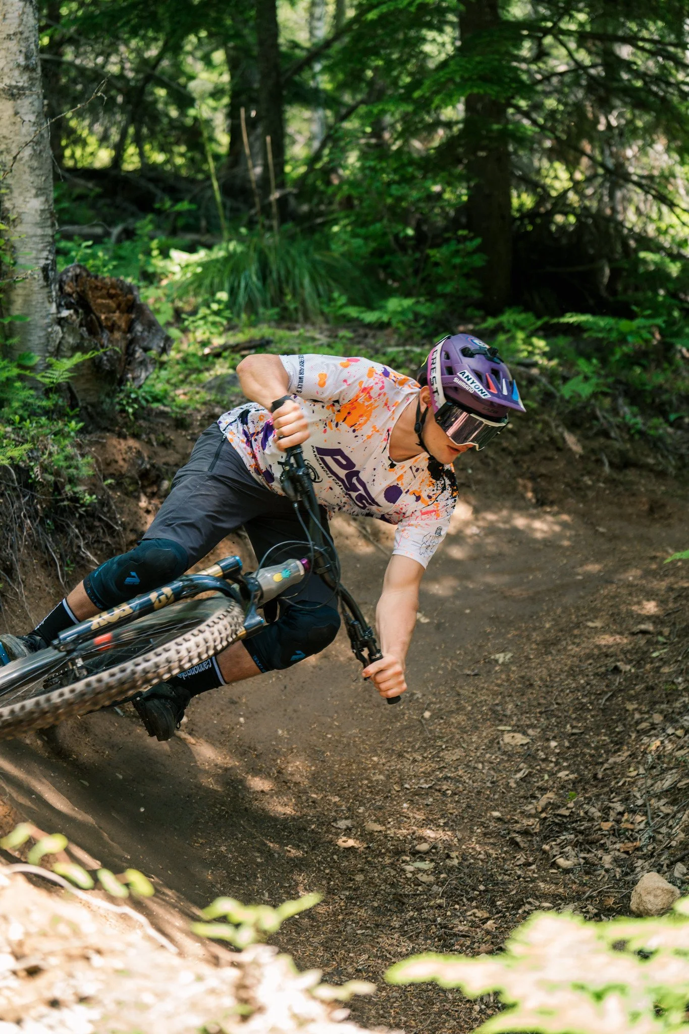 A mountain biker wearing a purple helmet, sunglasses, and a white jersey with orange and purple paint splashes, riding downhill on a dirt trail through a forest with green trees and foliage.