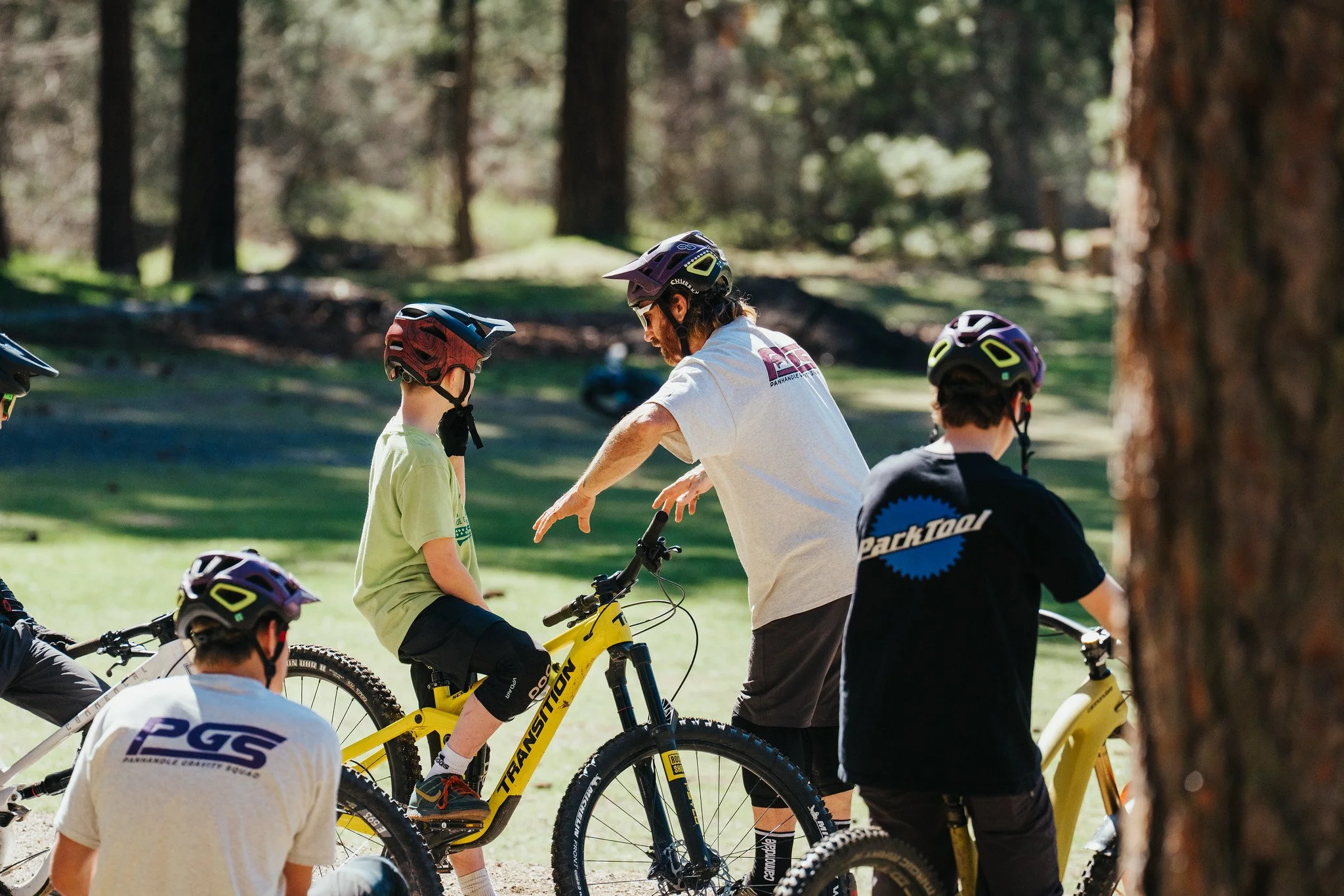 A group of children and an adult preparing for a mountain biking activity in a forested area, with some children sitting and others standing with their bikes, all wearing helmets.
