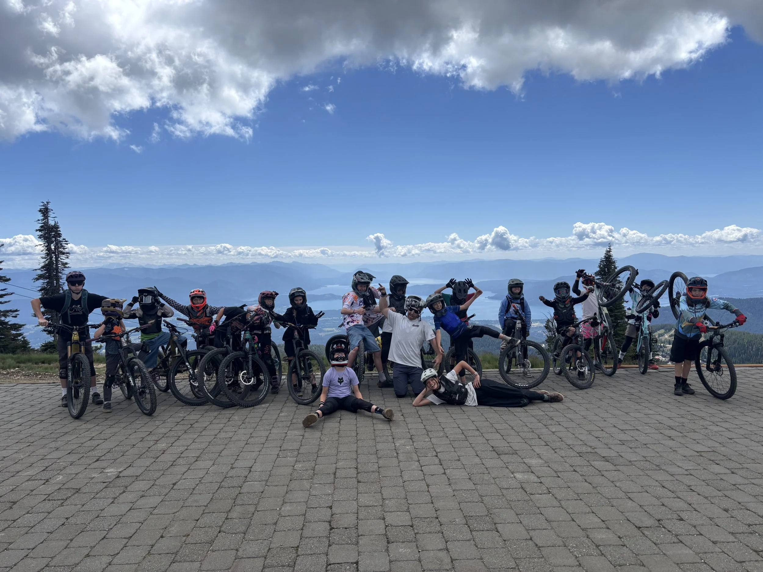 A group of children and teenagers with mountain bikes pose on a paved overlook with a scenic mountain view, some raising their bikes and making peace signs, under partly cloudy skies.