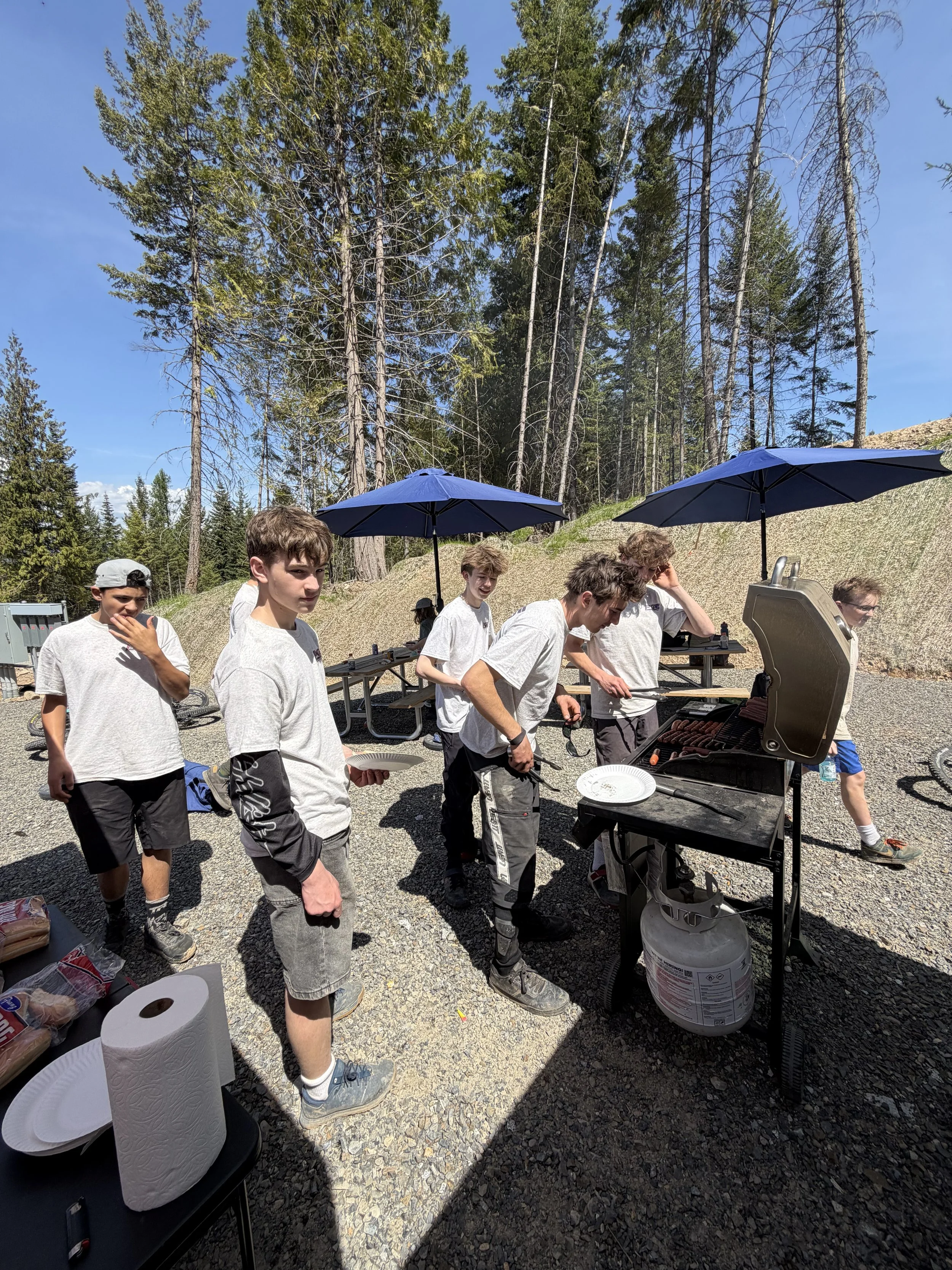 A group of young people gathered outdoors in a forested area, standing around a barbecue grill cooking meat, with some holding plates and utensils. They are wearing casual clothes and appear to be enjoying a cookout on a sunny day.