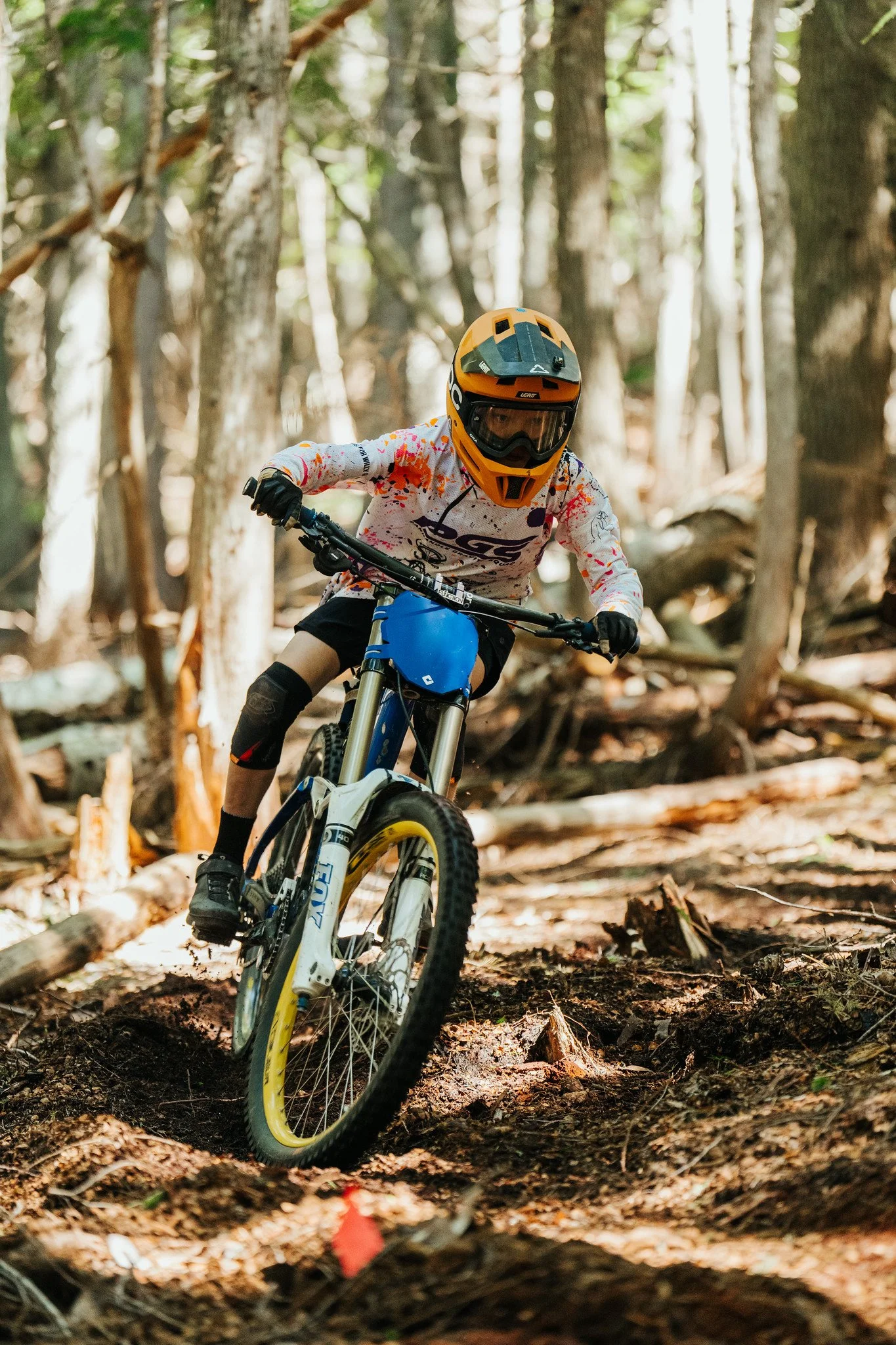 A person riding a mountain bike through a forest trail, wearing a yellow helmet, goggles, and a white jersey with red and purple accents.