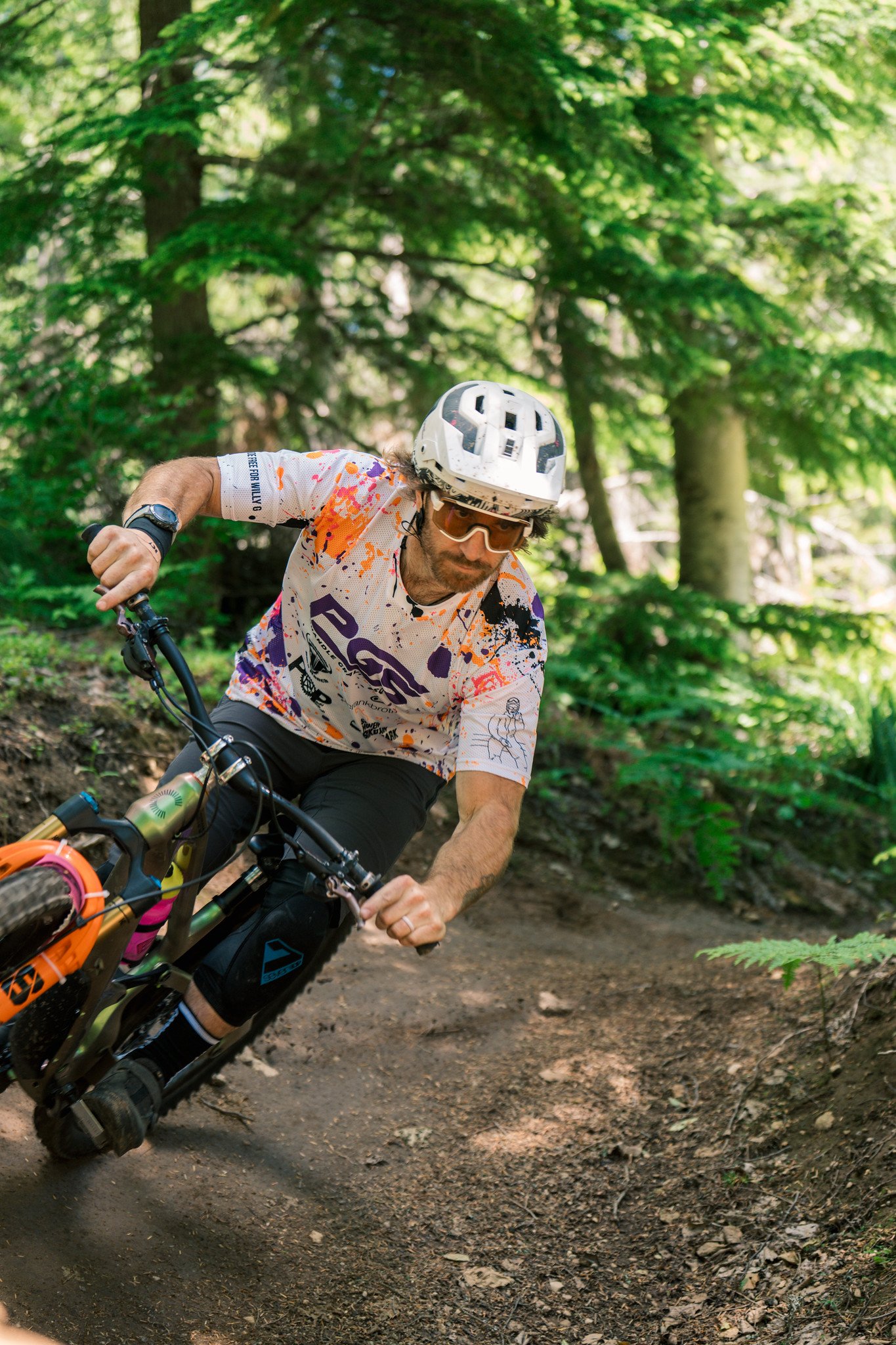 A man mountain biking on a forest trail, wearing a helmet, sunglasses, and a colorful jersey, surrounded by green trees and sunlight filtering through the leaves.