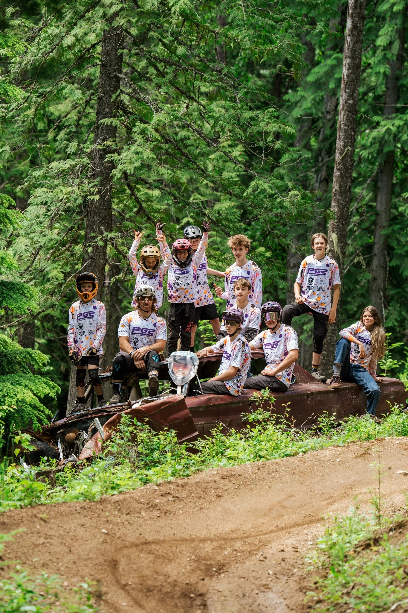 Teenagers and children wearing helmets and sports jerseys riding or sitting on a rusted, overturned car in a forested area with tall green trees and dirt trails.