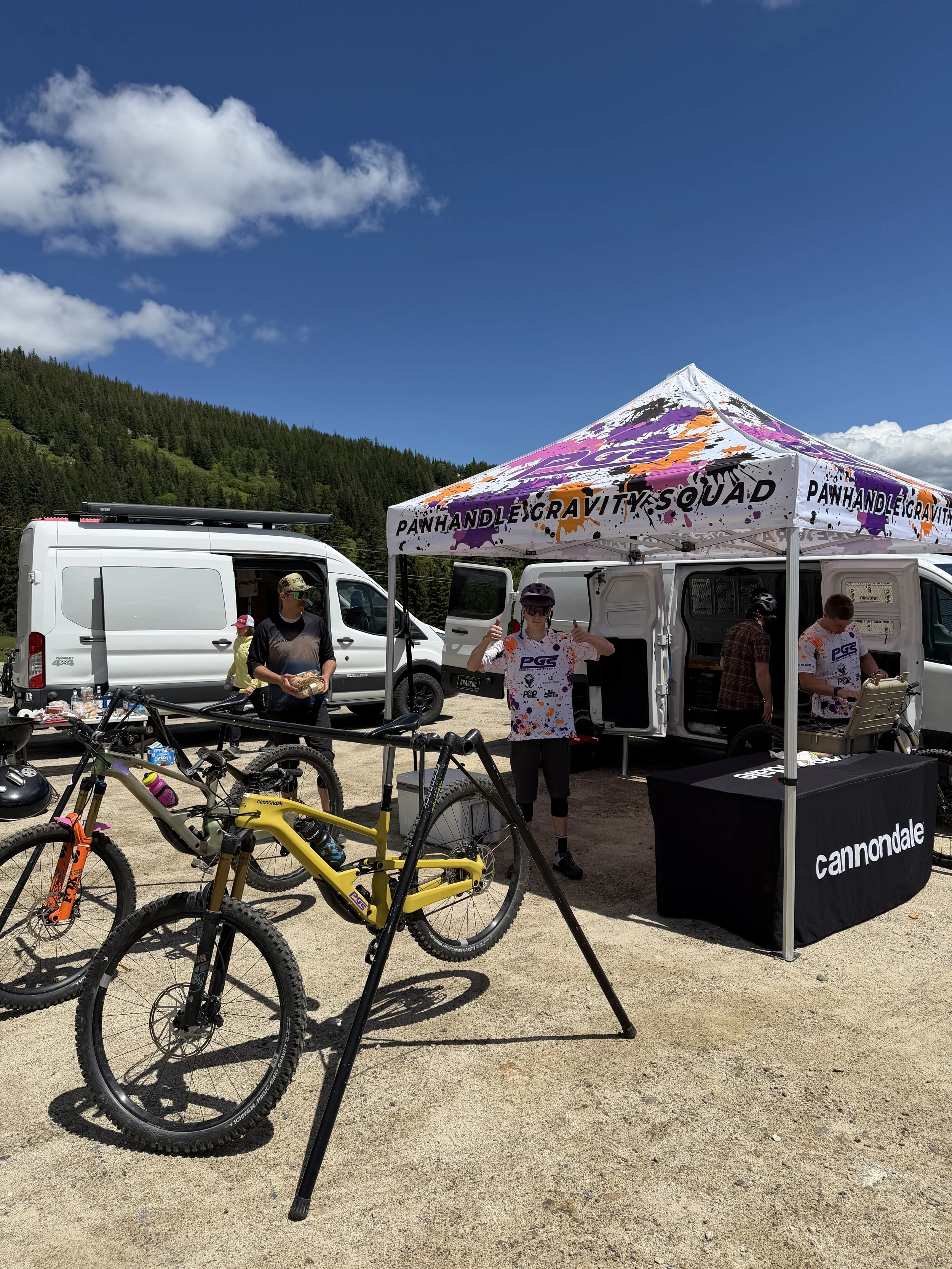 An outdoor scene showing a mountain bike, a white van, a canopy tent with the words 'Panhandle Gravity Squad' and 'Cannondale' on it, and several people under the tent. There are green mountains and a blue sky with clouds in the background.