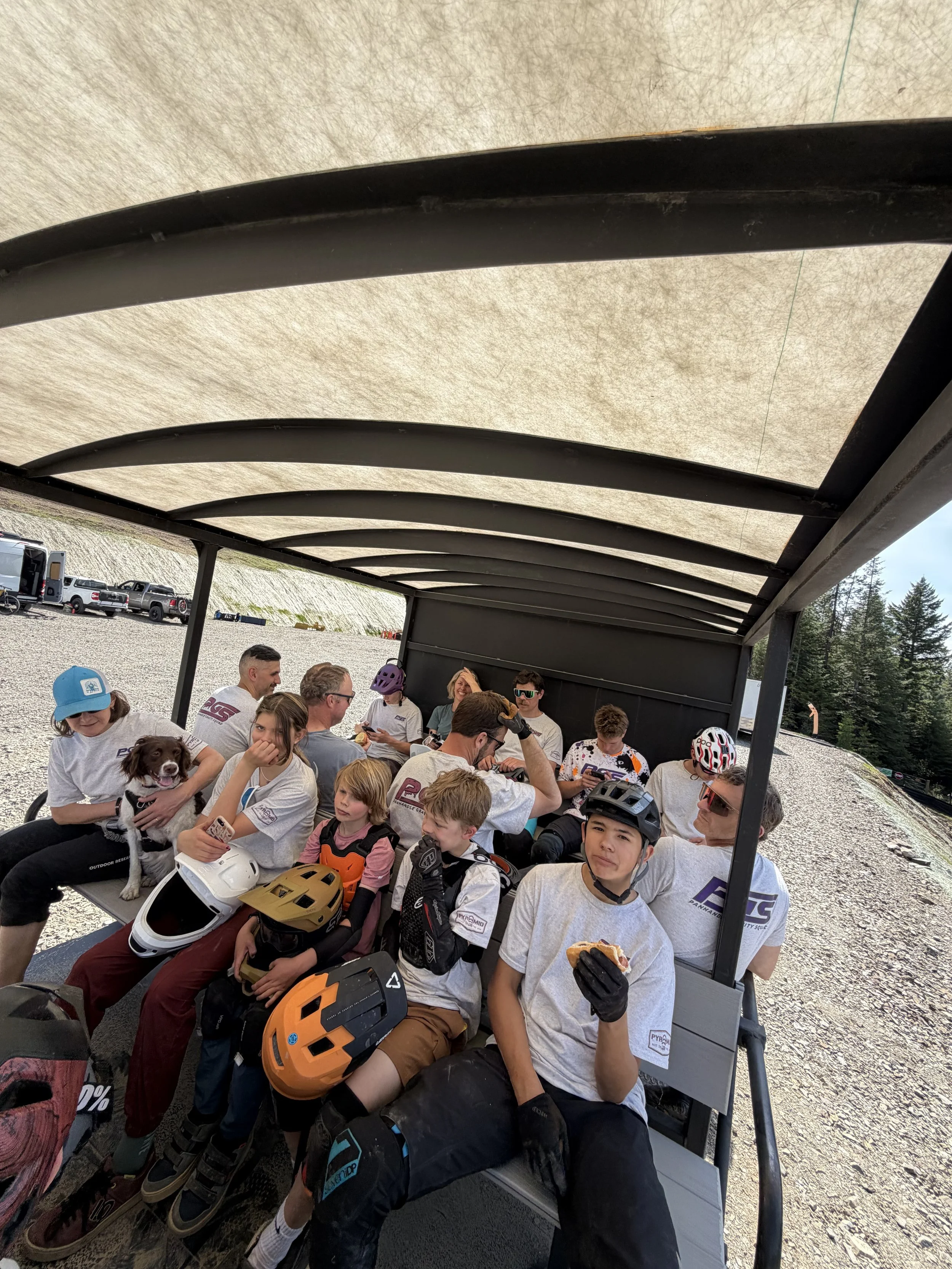 A group of children and adults sitting on a covered cart or tram, many with helmets and gloves, some holding food, in an outdoor setting with trees and parked trucks in the background.