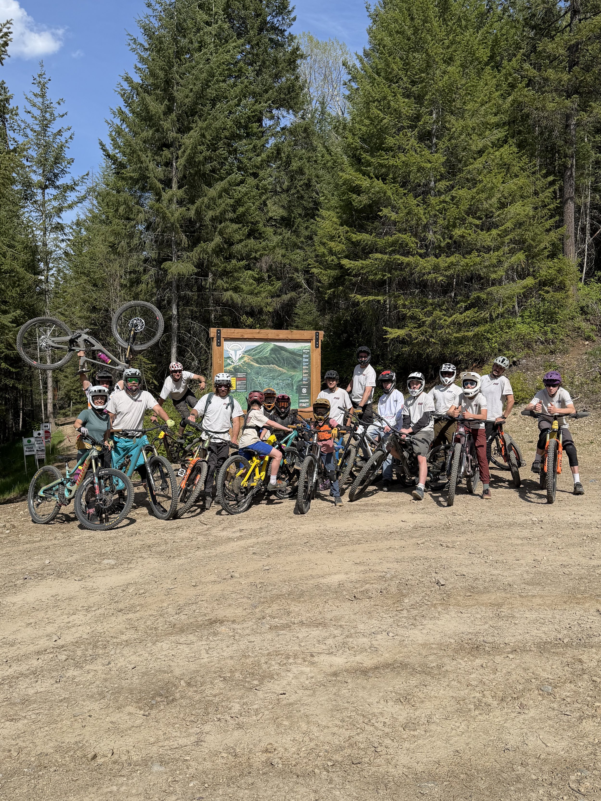 Group of children and adults posing with mountain bikes in a wooded outdoor area with a signboard behind them.