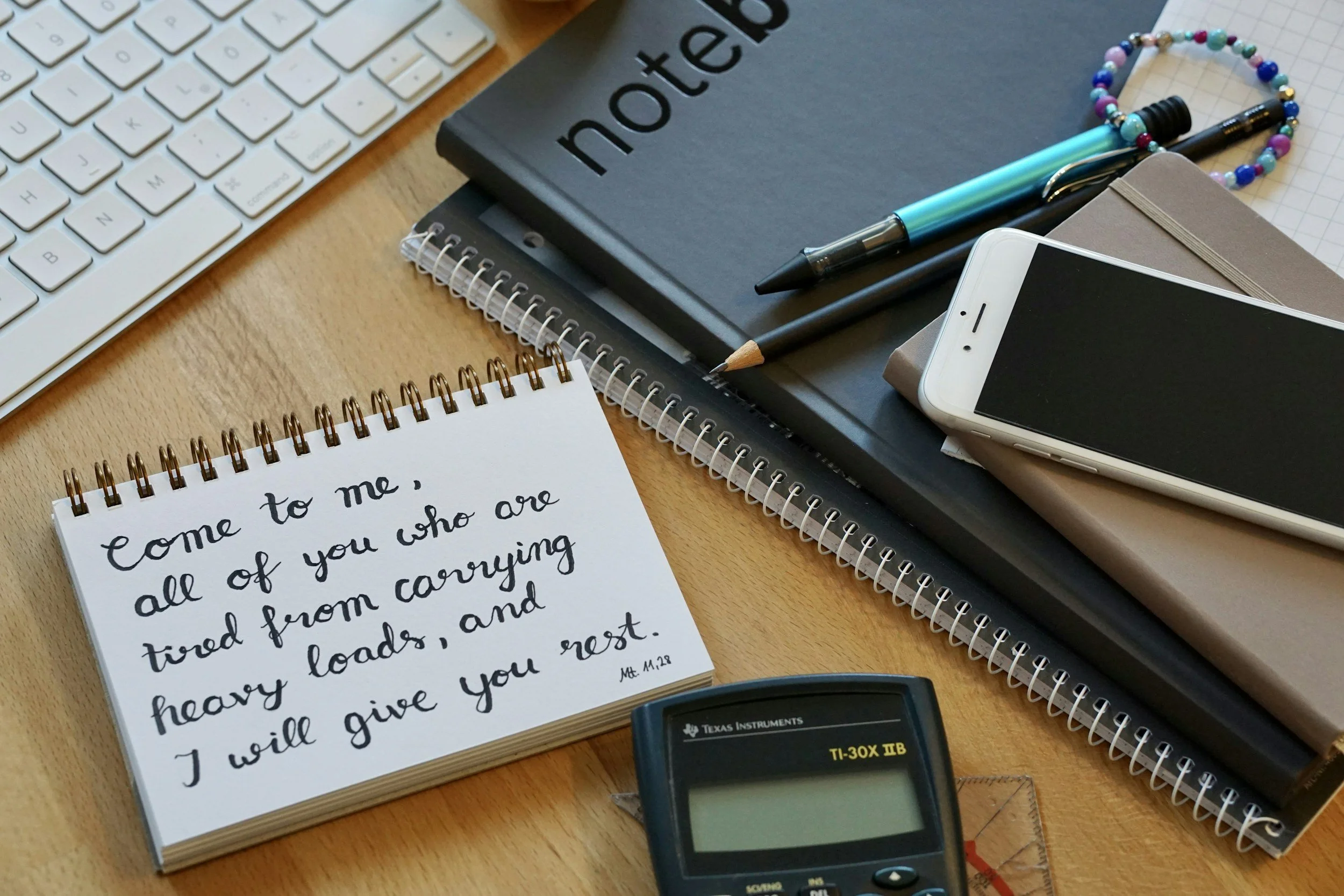 A desk with a computer keyboard, notebooks, pens, a beaded bracelet, a calculator, and a handwritten note with a Bible verse.