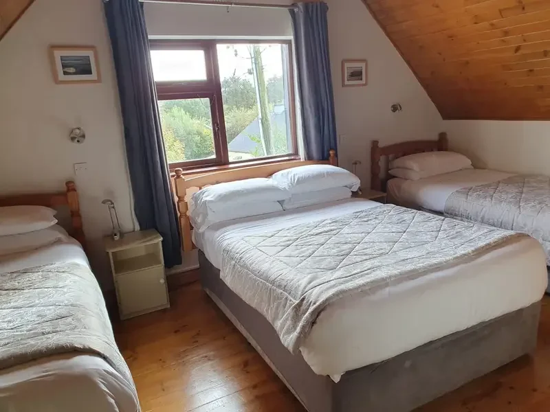 A cozy bedroom with three beds, one large bed in the center and two smaller beds on each side, all with white bedding. A window with blue curtains shows trees outside. The room has wooden floors and a slanted wooden ceiling. Bedrooms for four people.