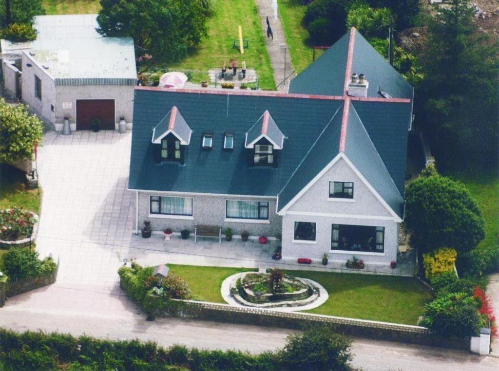 Aerial view of a large house with blue roof, white walls, front garden with a circular flower bed, and driveway. Small backyard with picnic table, umbrella, trampoline, hammocks and lawn. Bike storage and private car park. Self check in option.