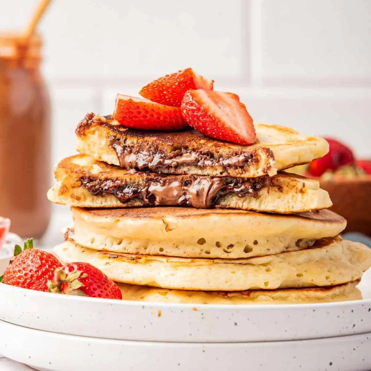 Stack of four pancakes topped with sliced strawberries and chocolate syrup, on a white plate with additional strawberries on the side.