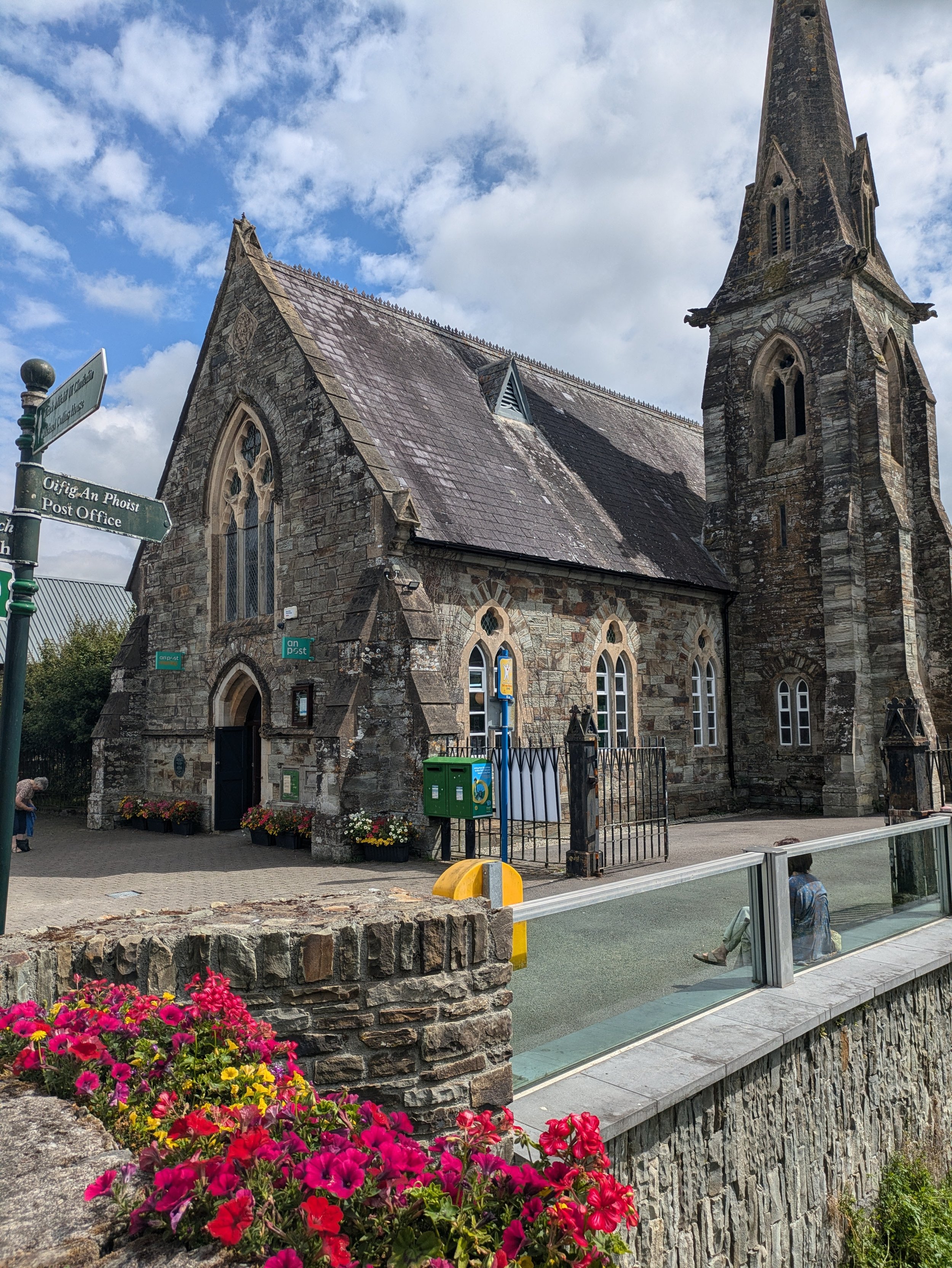 A stone church with a tall steeple and pointed arch windows, located on a street corner with street signs, colorful flowers in the foreground, and a partly cloudy sky overhead. Post office that has been converted from an old church.