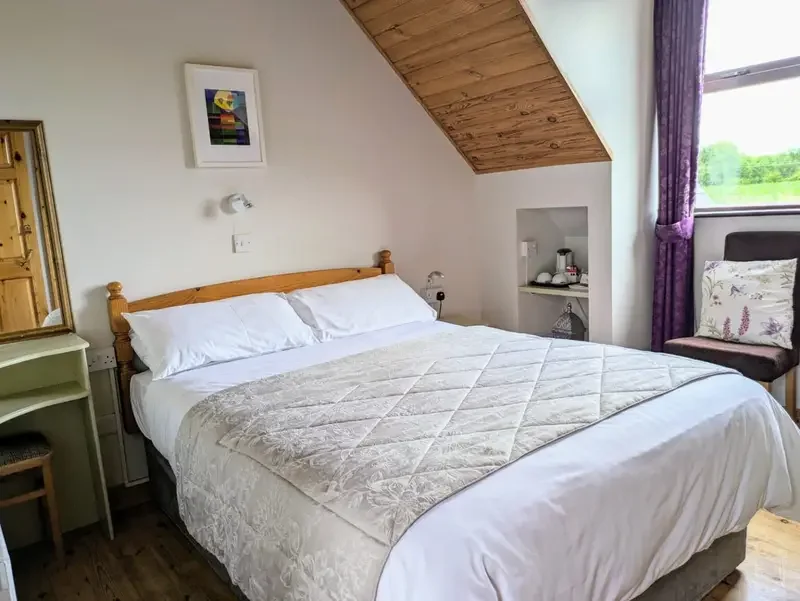 A cozy bedroom with a sloped wooden ceiling and a large bed with white linens and a beige quilted coverlet. There is a small green bedside table with a mirror, a framed colorful artwork on the wall, and a window with purple curtains revealing a green landscape outside. A black chair with a floral pillow is near the window, and there is a small alcove with a coffee maker and cups.