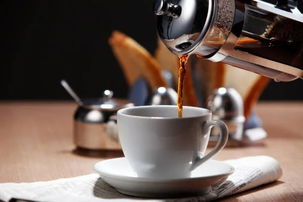 A white cup being filled with coffee from a french press coffee pot on a wooden table with toast in the background.