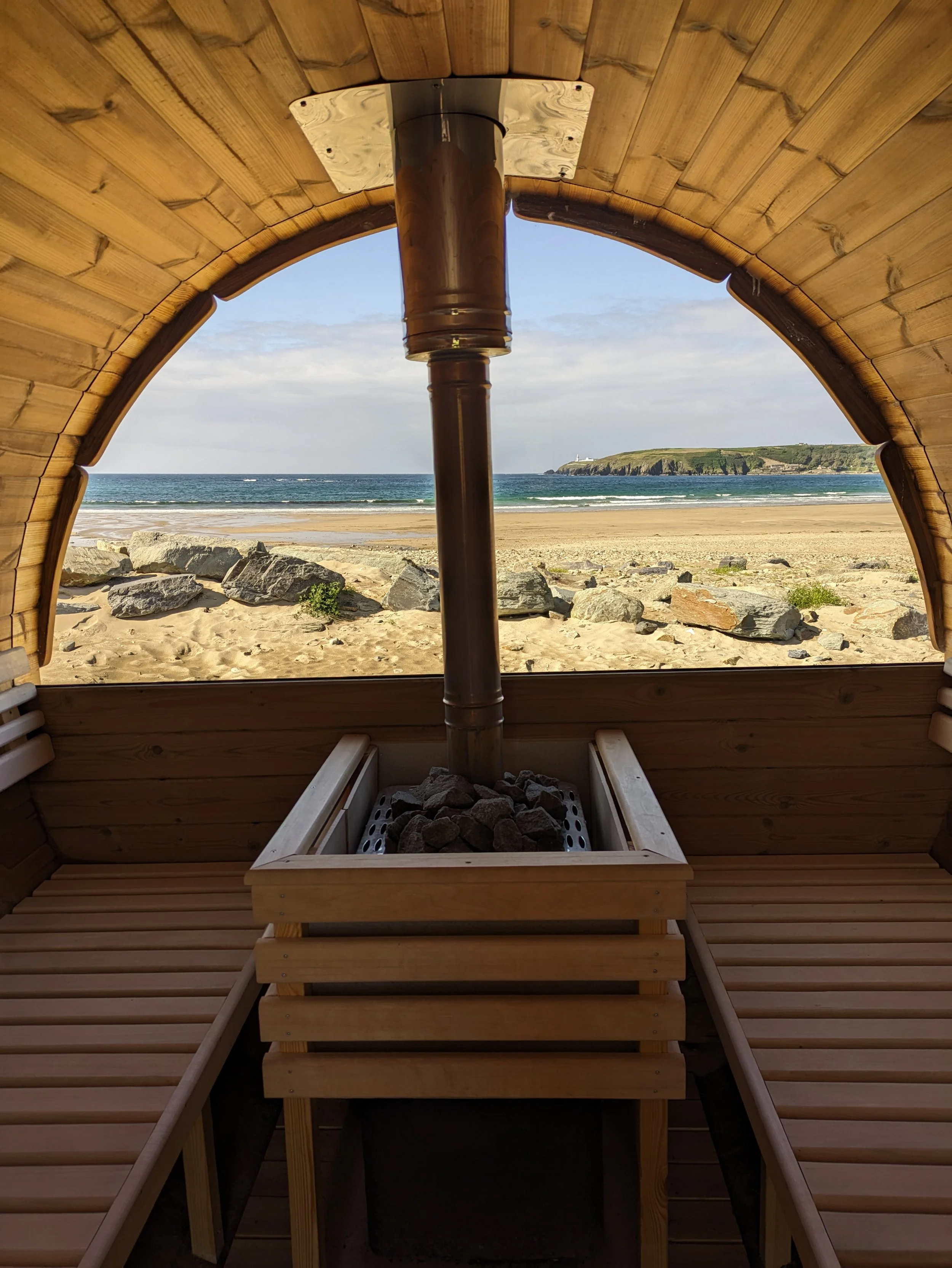 View from inside a wooden sauna looking out through an arched opening at a beach with rocks, the ocean, and a lighthouse on a distant hill. Redwood barrel sauna and ocean dipping.