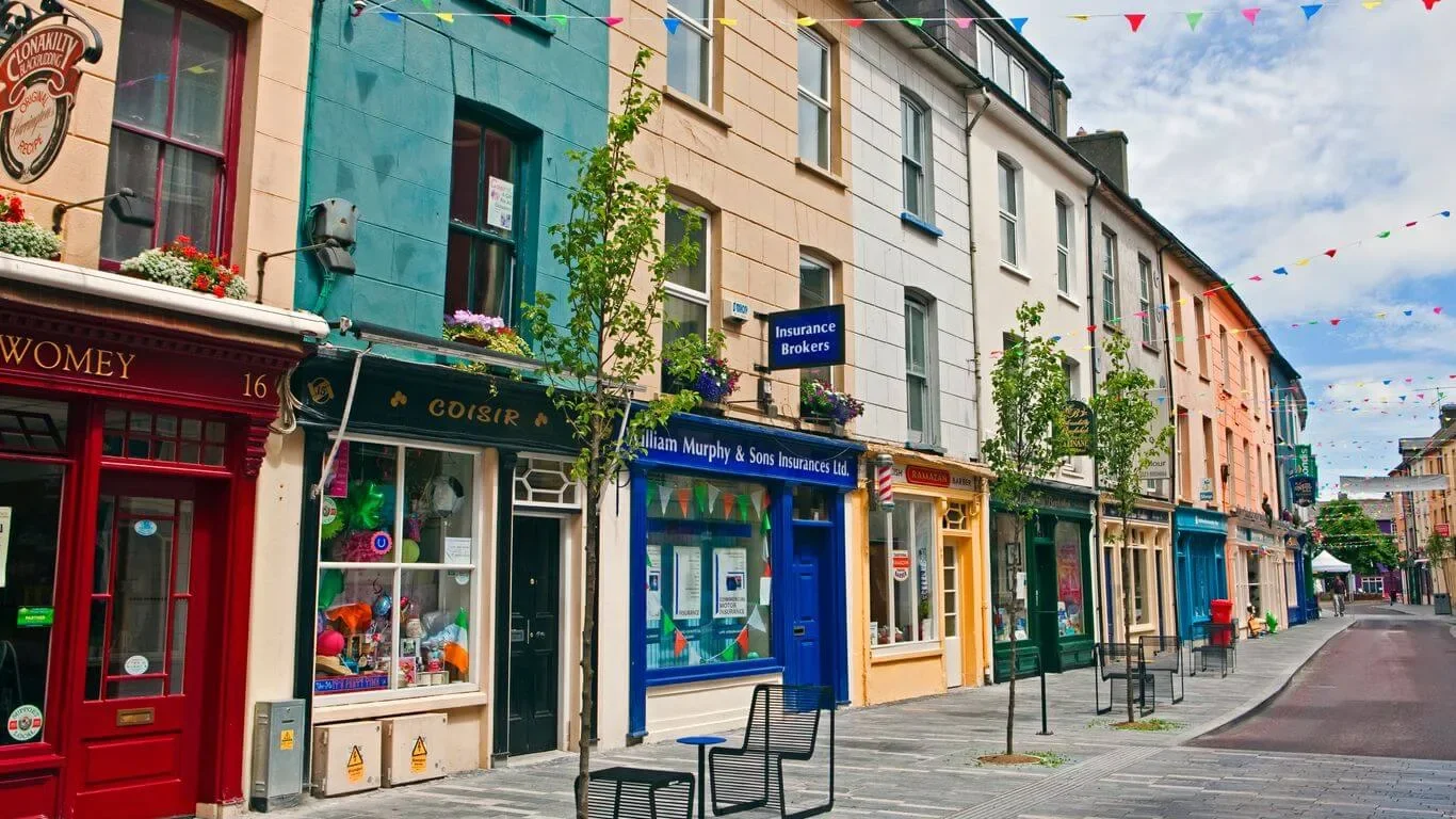 Colorful row of shops and businesses on a quiet street with trees, benches, and colorful bunting in the sky. Cultural town with lots of color and gorgeous shopfronts.