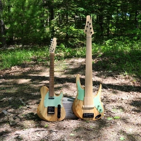 Two oak wooden electric bass guitars leaning against a small log in a forest setting with green trees and sunlight.