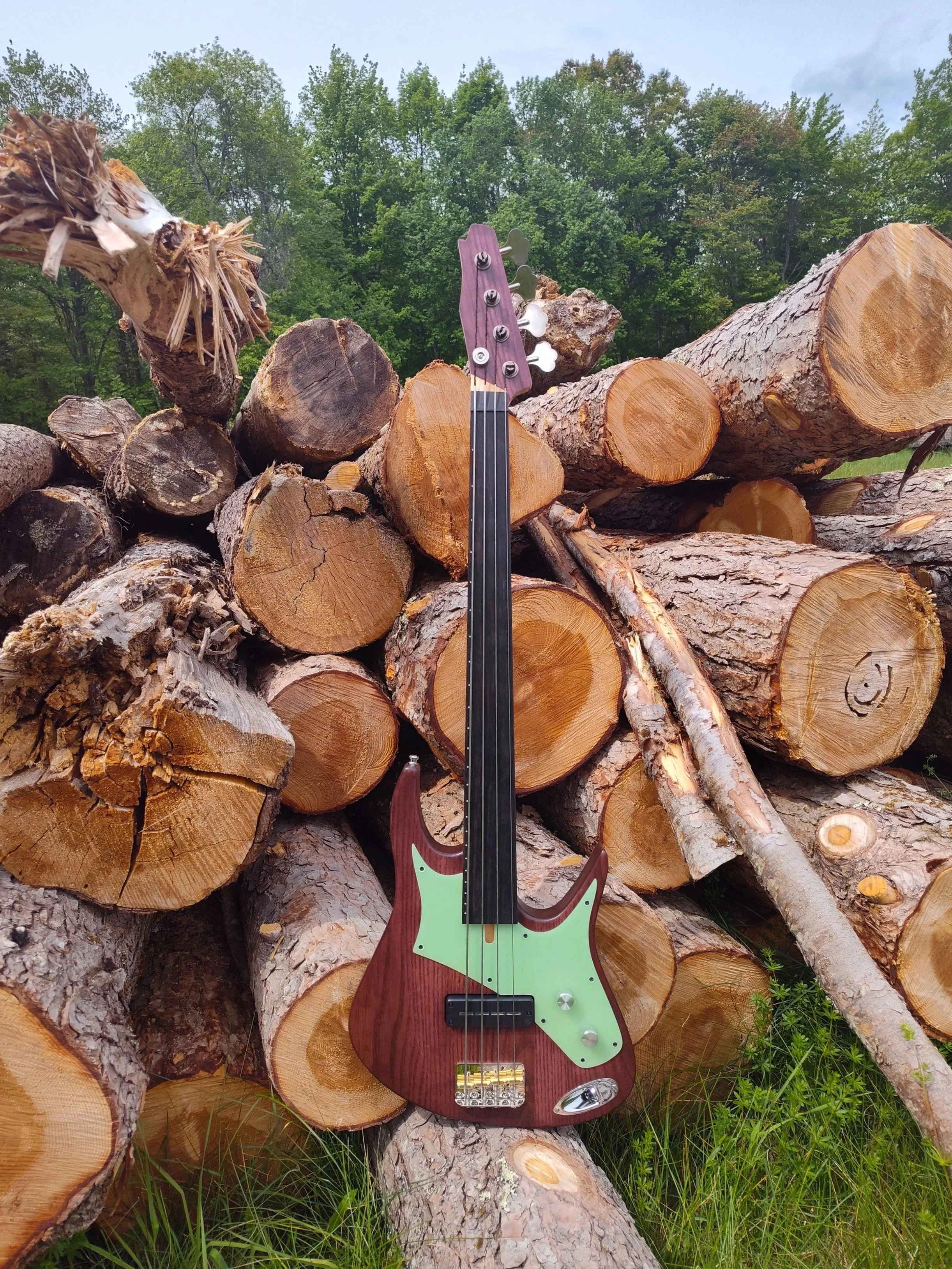 A mahogany guitar placed against a large pile of cut logs with a background of green trees and an overcast sky.