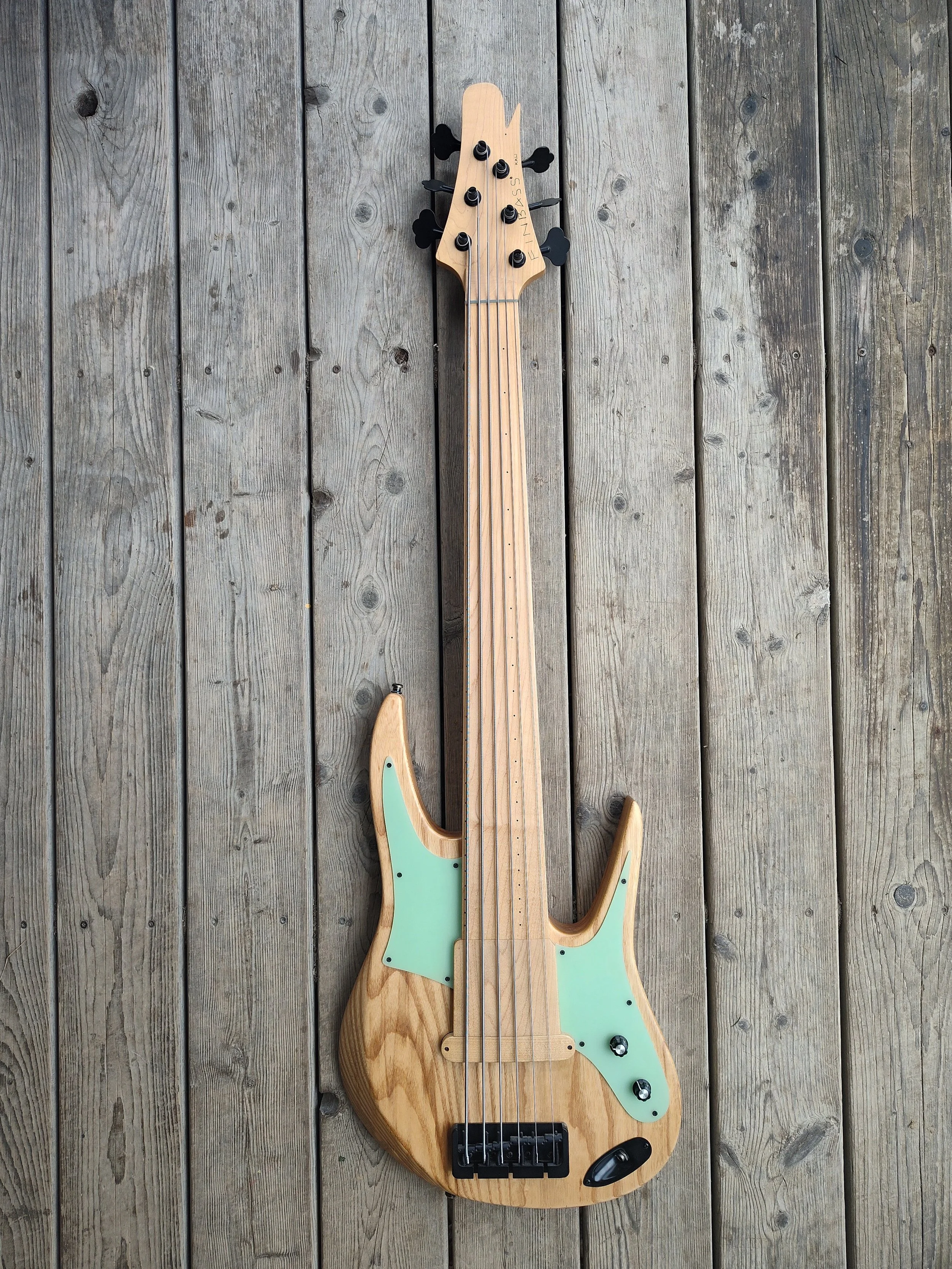 Electric bass guitar with a natural wood finish, black hardware, and mint green pickguard, lying on weathered wooden planks.