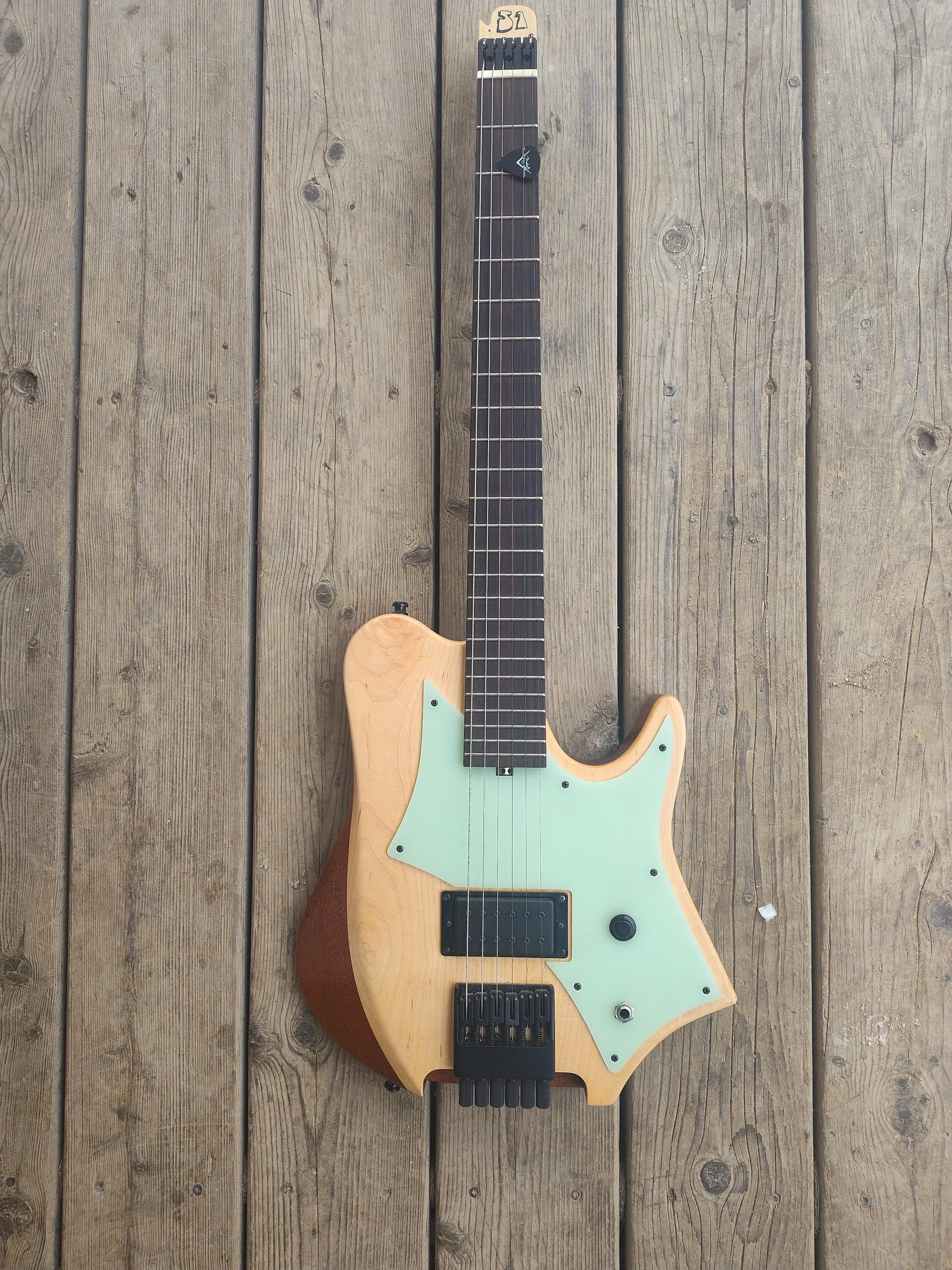 An electric guitar with a natural wood finish, black hardware, and a mint green pickguard lying on a wooden deck.