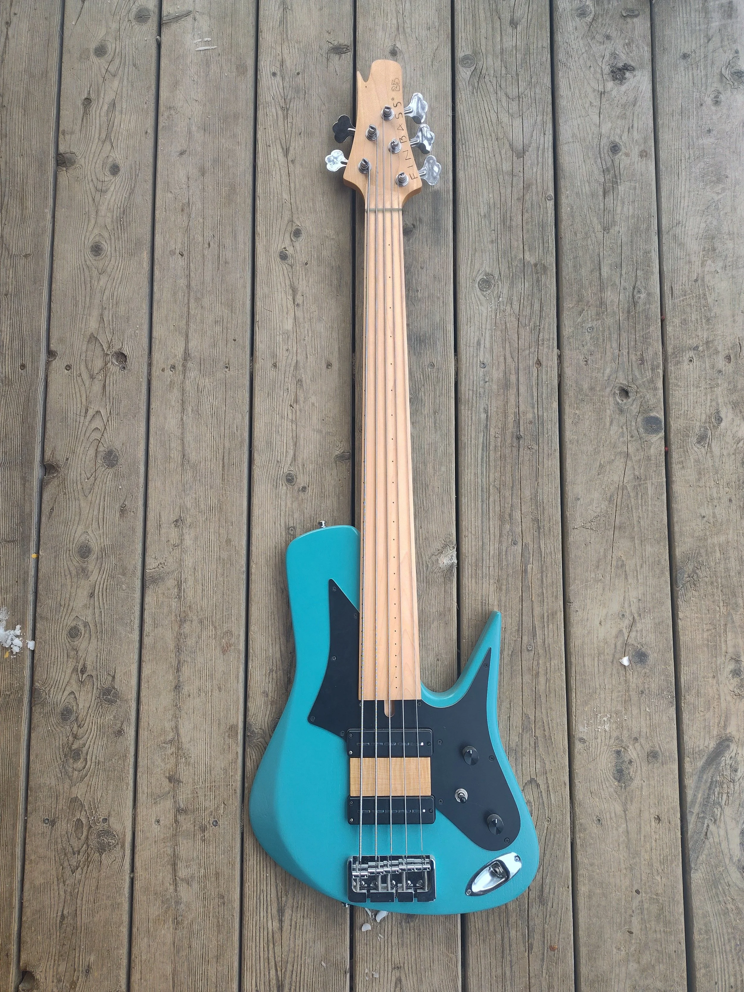 A blue electric bass guitar with a black pickguard is laying on a wooden deck. The guitar has four strings, a control knob, and a bridge at the bottom.