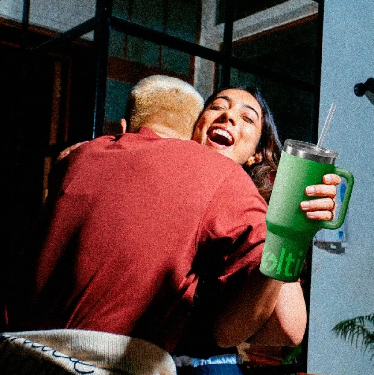 Dos personas abrazándose con una sonrisa, una de ellas sostiene un vaso con una bebida y una pajilla.