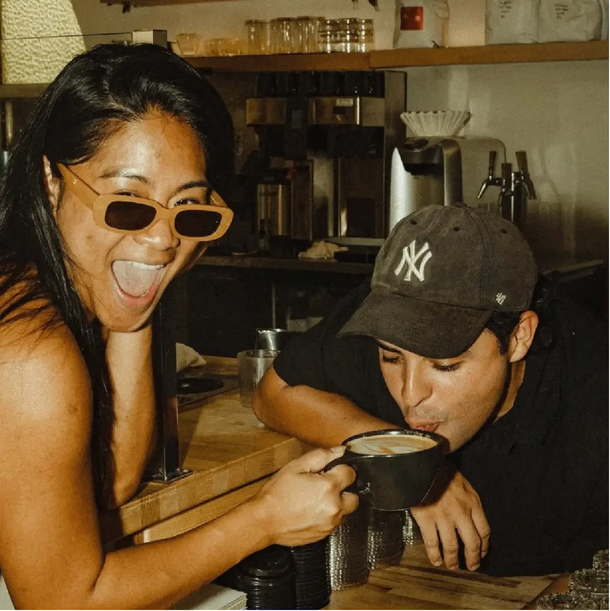 Dos personas en un ambiente de cafetería, una mujer feliz con gafas de sol amarillas sonriendo y sosteniendo una taza, y un hombre con gorra de béisbol bebiendo de la misma taza.
