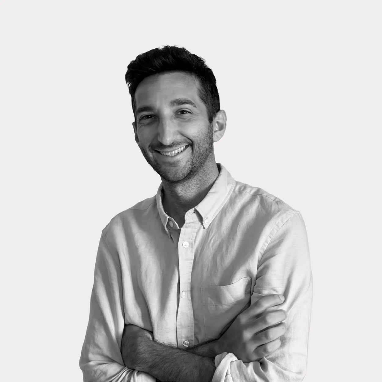 Black-and-white portrait of a smiling man with his arms crossed, wearing a slightly wrinkled long-sleeved shirt, against a white background.