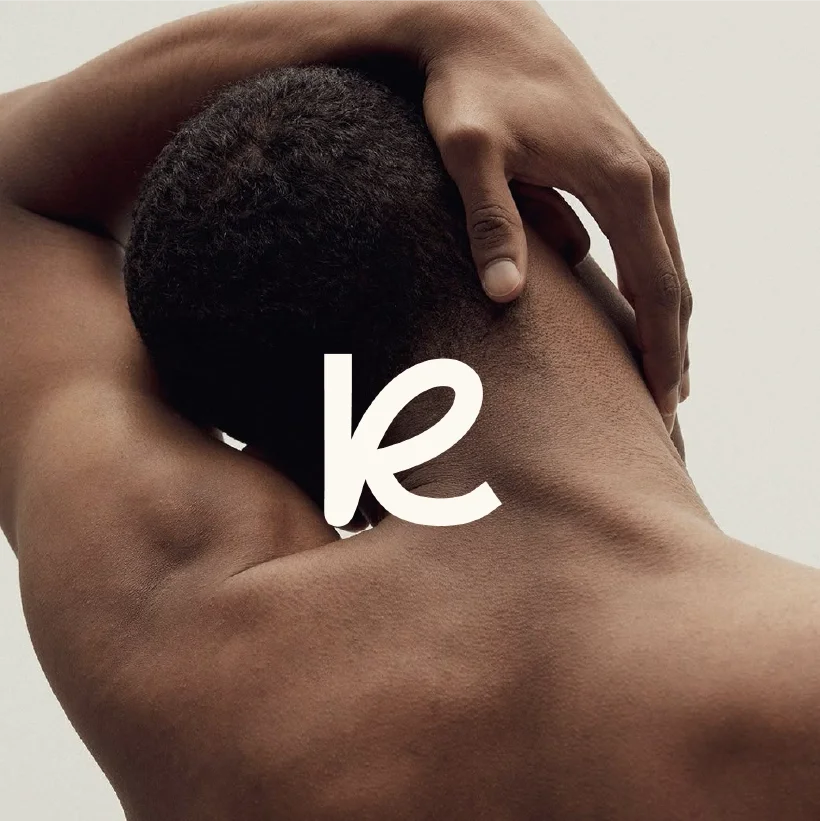 A nude person with their back and neck visible, touching their head with both hands, against a neutral background.