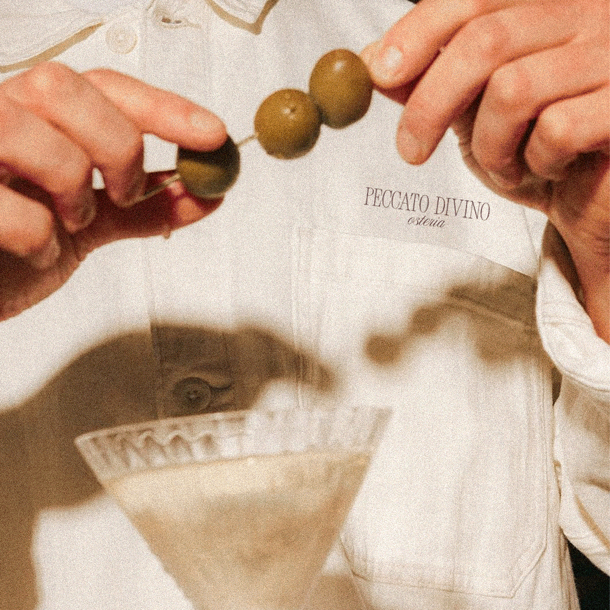 A person holding a skewer with olives and a paper bag with the words "PECATO DIVINO" written in cursive.