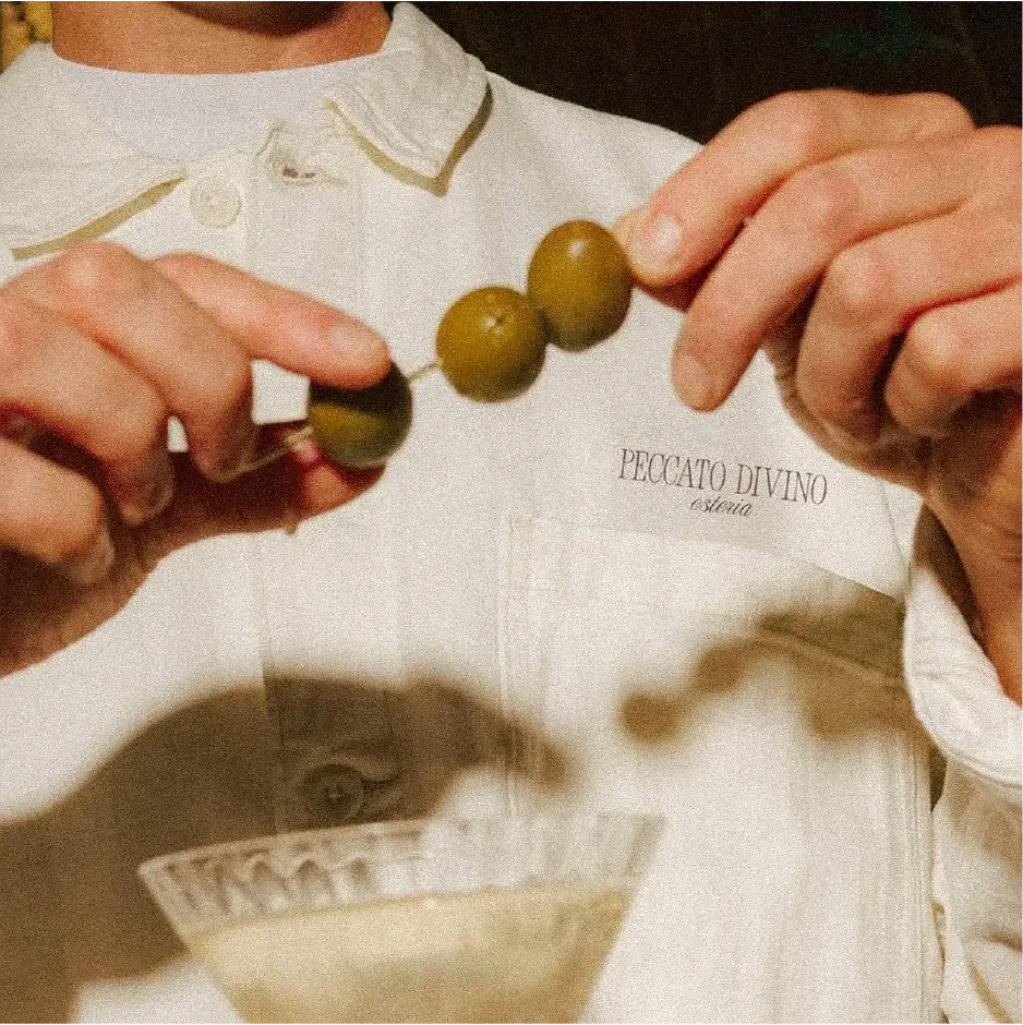 A person holding a rosary with three green beads, wearing white clothing, and a name tag that reads “Peccato Divino Estetica.”