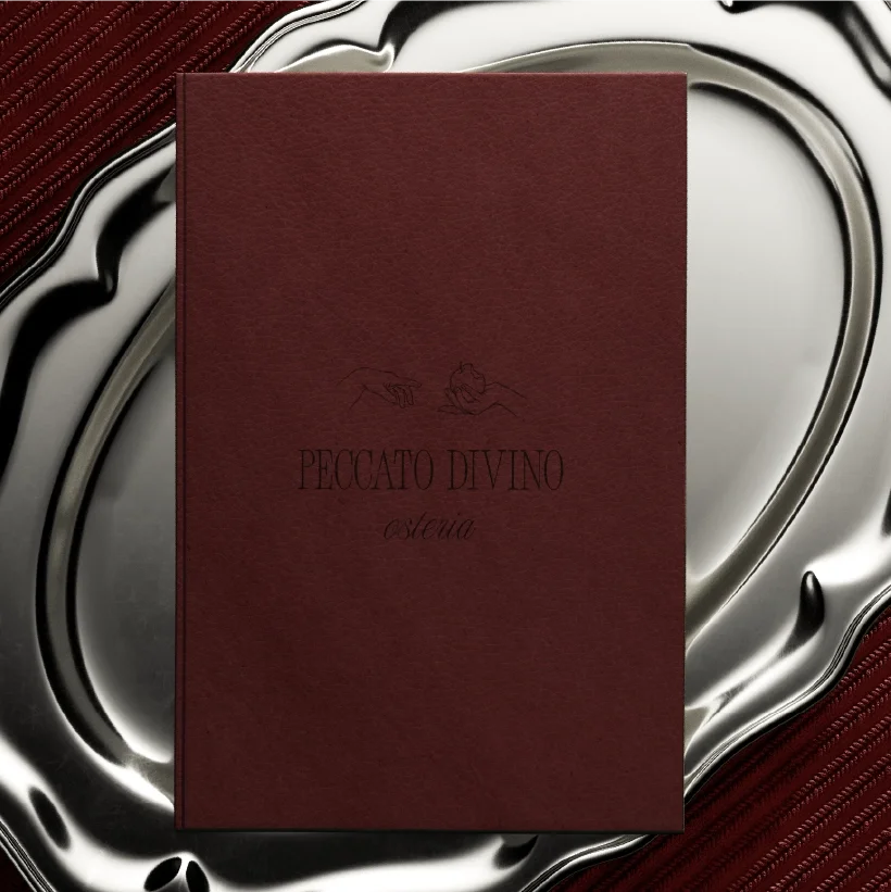 A book with a red cover, titled “Divine Sin,” featuring a drawing of two hands—one outstretched and the other in a prayer position—against a background of a silver table and red fabric.