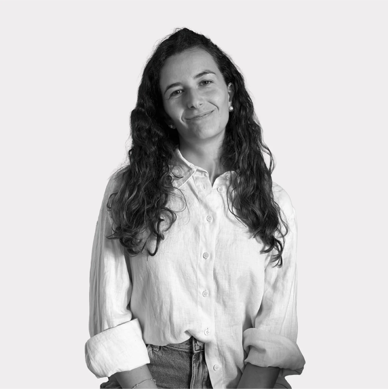 Black-and-white portrait of a woman with curly hair, smiling, wearing a white shirt and jeans, against a light gray background.