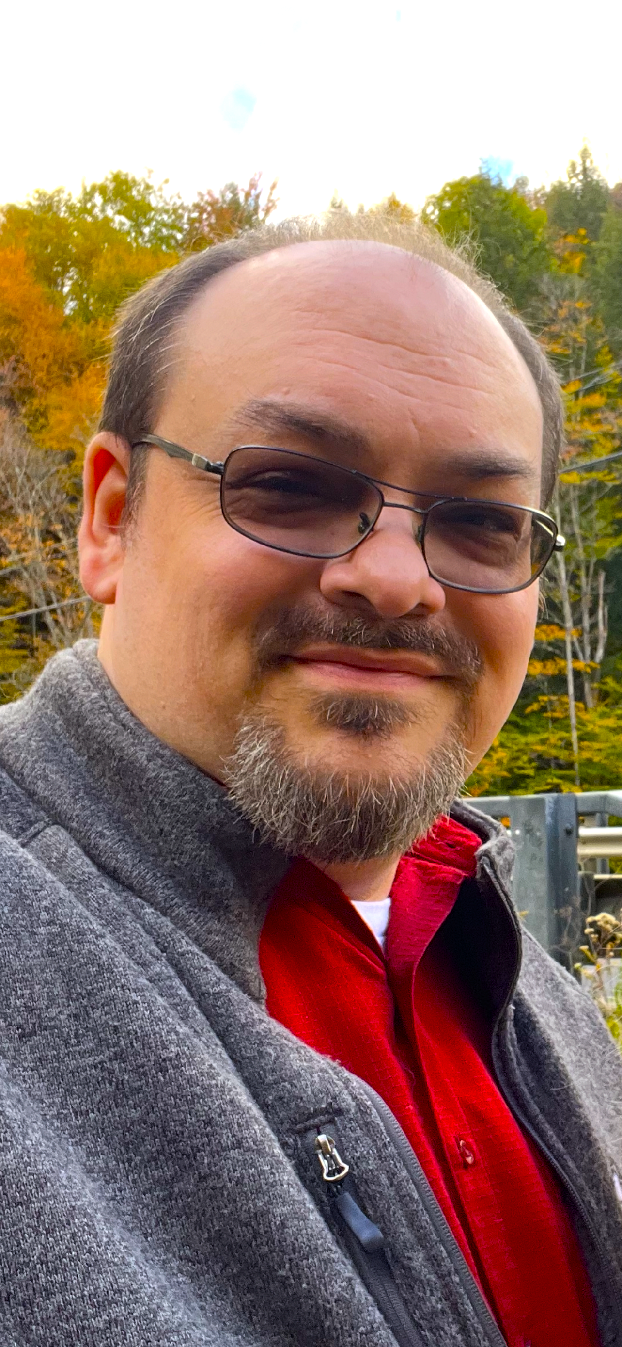 Billy Poe wearing glasses, a gray jacket, and a red shirt outdoors with fall foliage in the background.