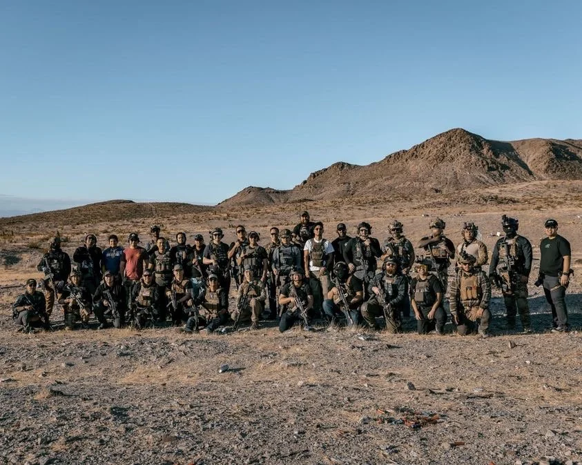 Group of soldiers in tactical gear standing and kneeling on a desert terrain with mountains in the background.
