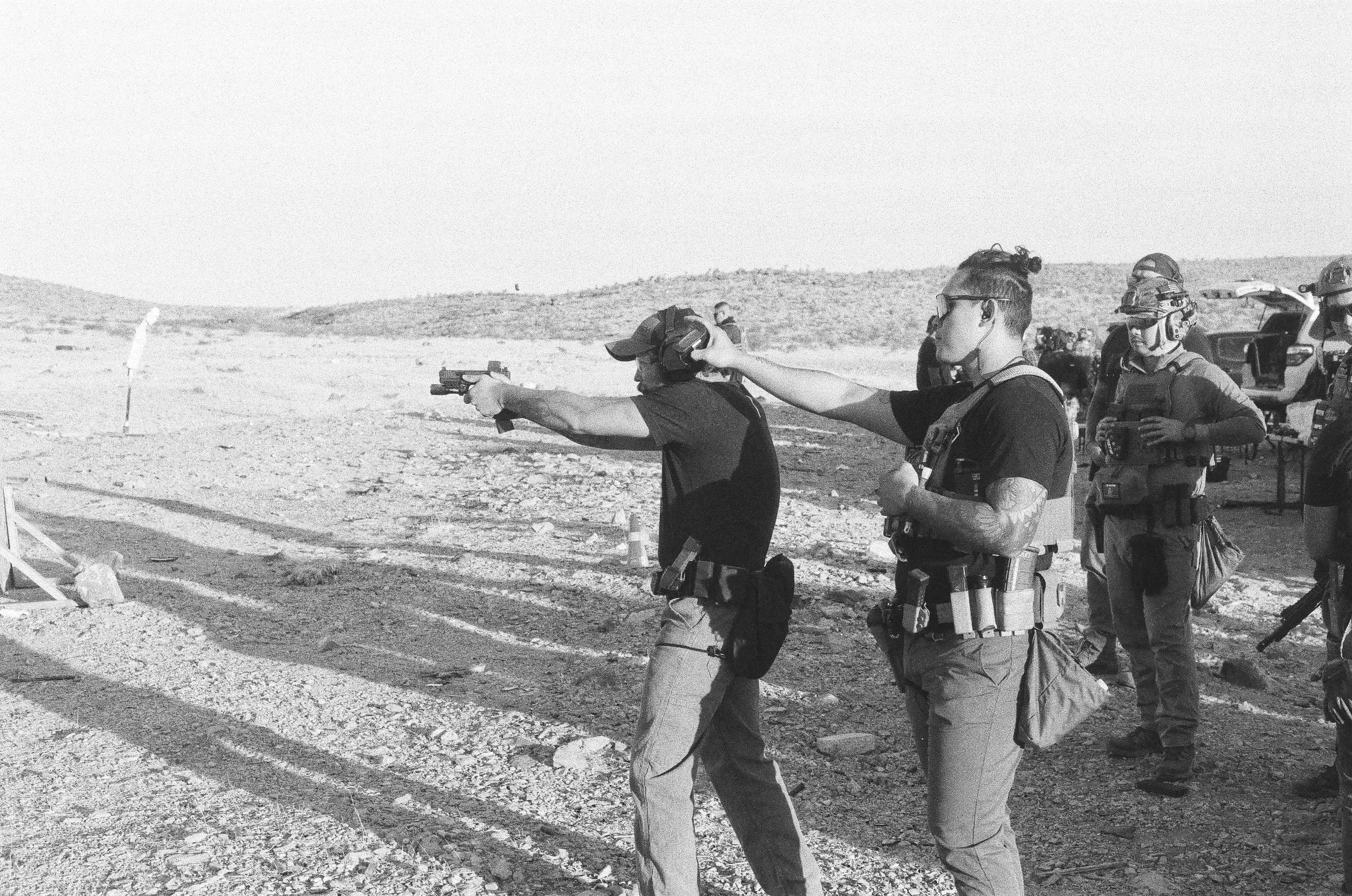 A group of armed individuals practicing shooting at an outdoor shooting range in a desert landscape. They are wearing protective gear and tactical clothing, aiming firearms.